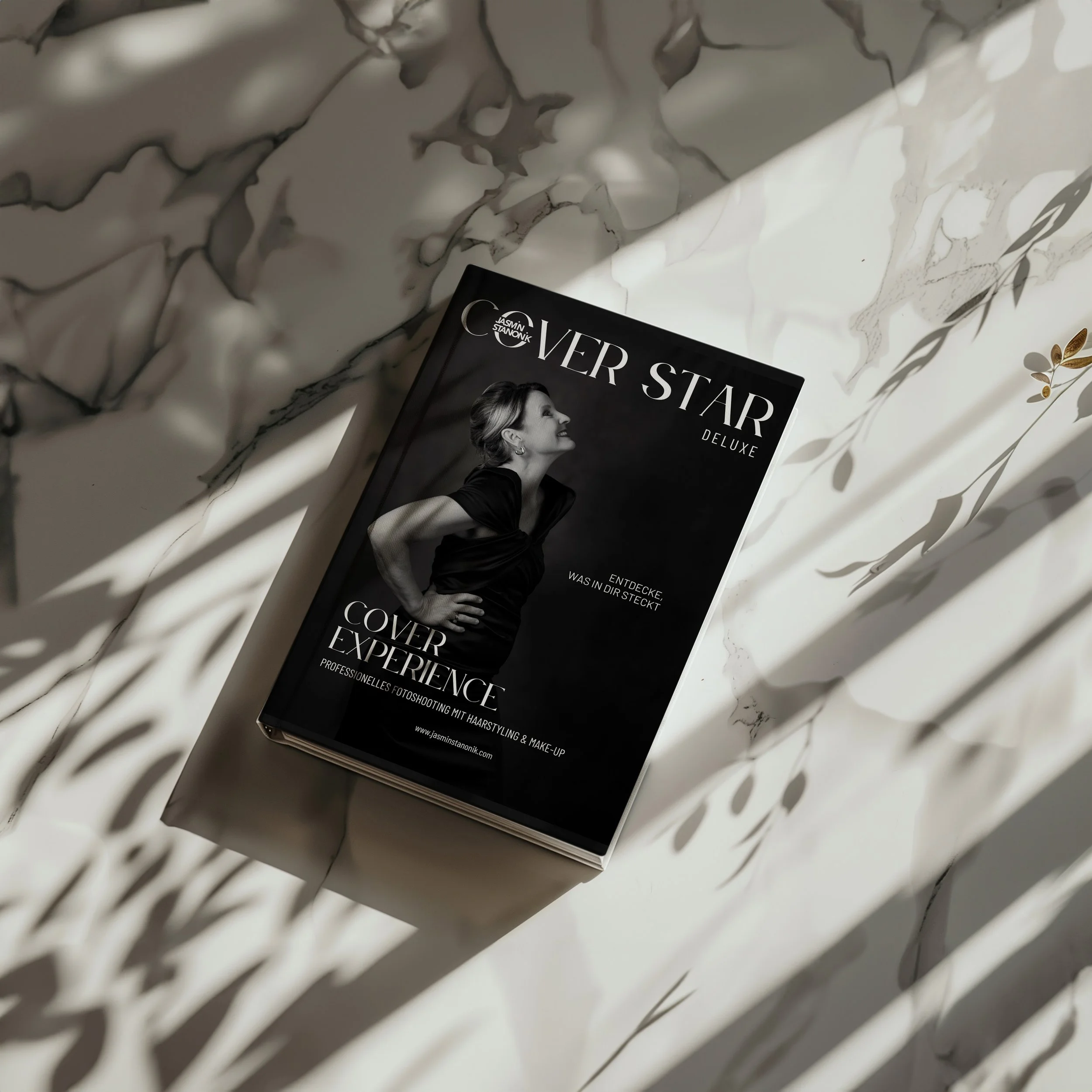 Ein schwarzes Magazin namens 'COVER STAR DELUXE' liegt auf einem weißen Marmor-Tisch, umgeben von Schatten von Blättern, mit einem Bild eines lächelnden, elegant gekleideten Frauen in Schwarz auf dem Cover.