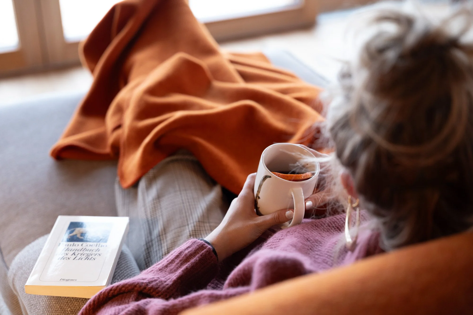 Frau liegt auf einem Sofa, trinkt Tee und liest ein Buch.
