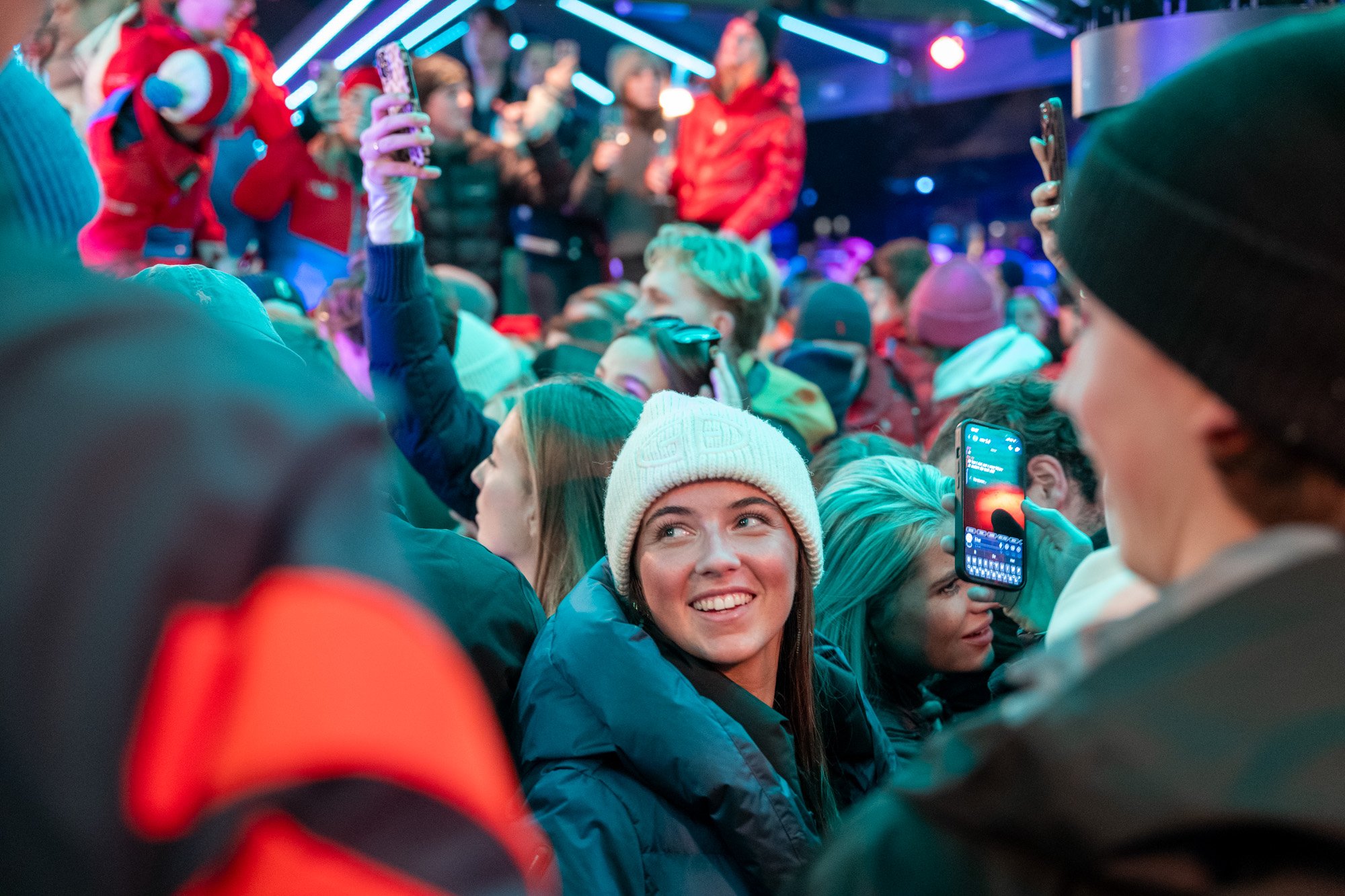 Junge Frau mit weißer Mütze lächelt unter einer Meute von Menschen in einem Nachtclub mit bunten Lichtern, während sie ein Smartphone in die Kamera hält.