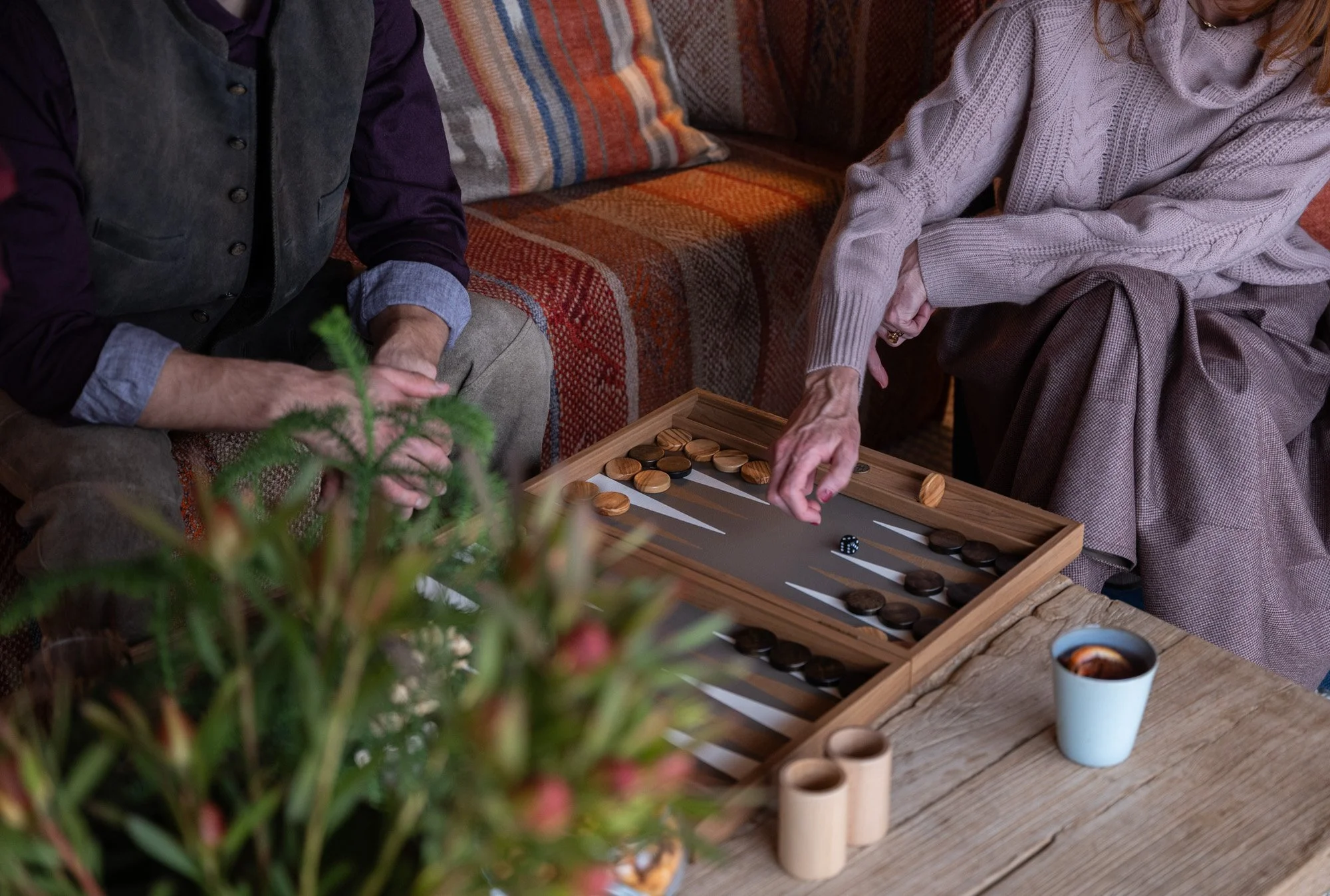 Zwei Personen spielen Backgammon an einem Holztisch, während sie eine Tasse Kaffee trinken. Im Vordergrund sind Pflanzen auf dem Tisch sichtbar.