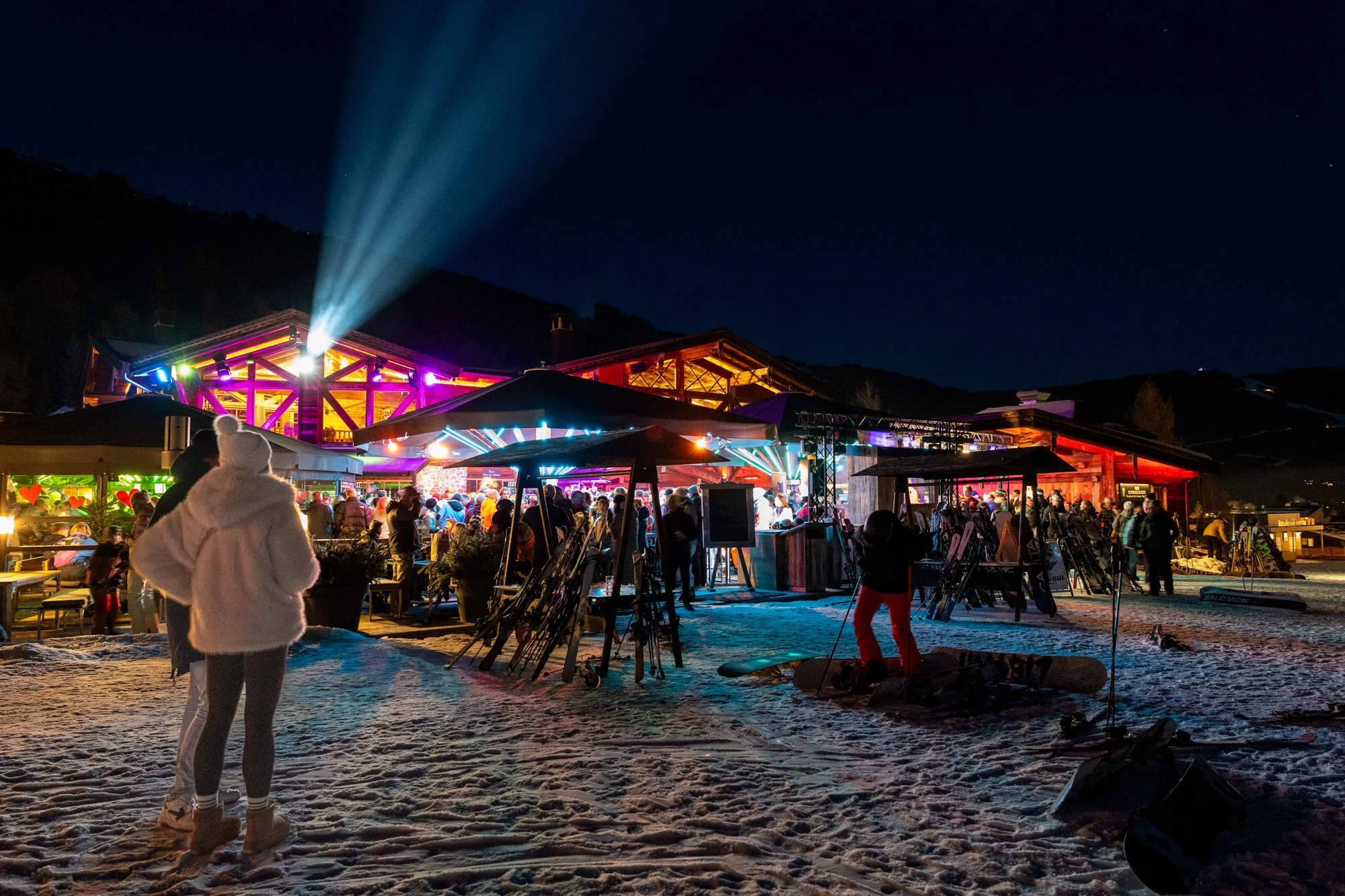 Menschen feiern auf einer verschneiten Skihütte bei Nacht mit bunten Lichtern und Skiern im Vordergrund.