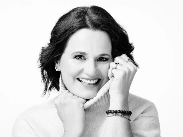 Schwarz-weiß-Foto einer lächelnden Frau, die ihre Hand an ihr Gesicht hält und in die Kamera schaut.