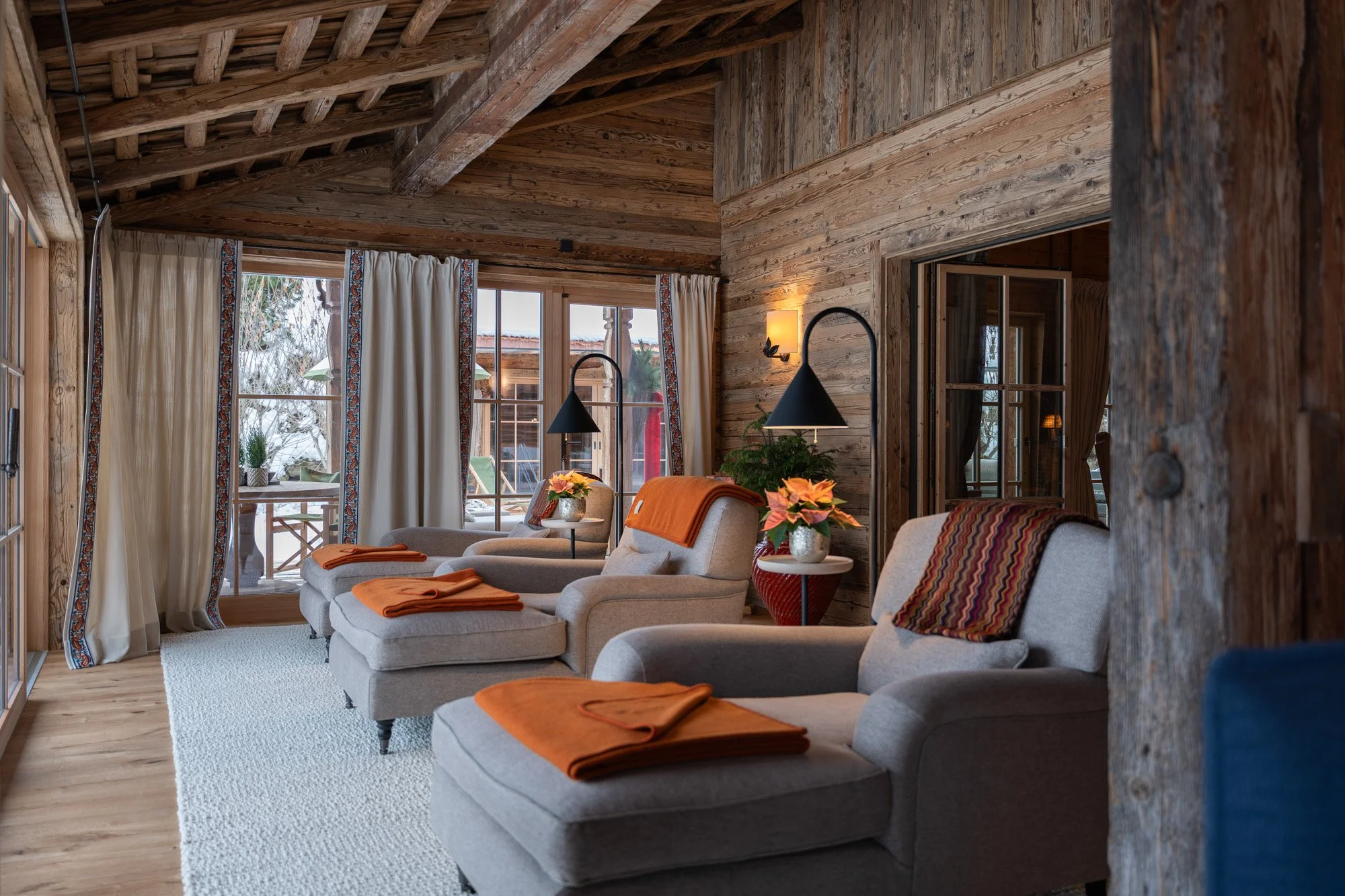 Gemütliches Wohnzimmer mit Holzdecken, großen Fenstern, bequemen Sessel, orange Decken und Tischdecken, sowie floralen Dekorationen.