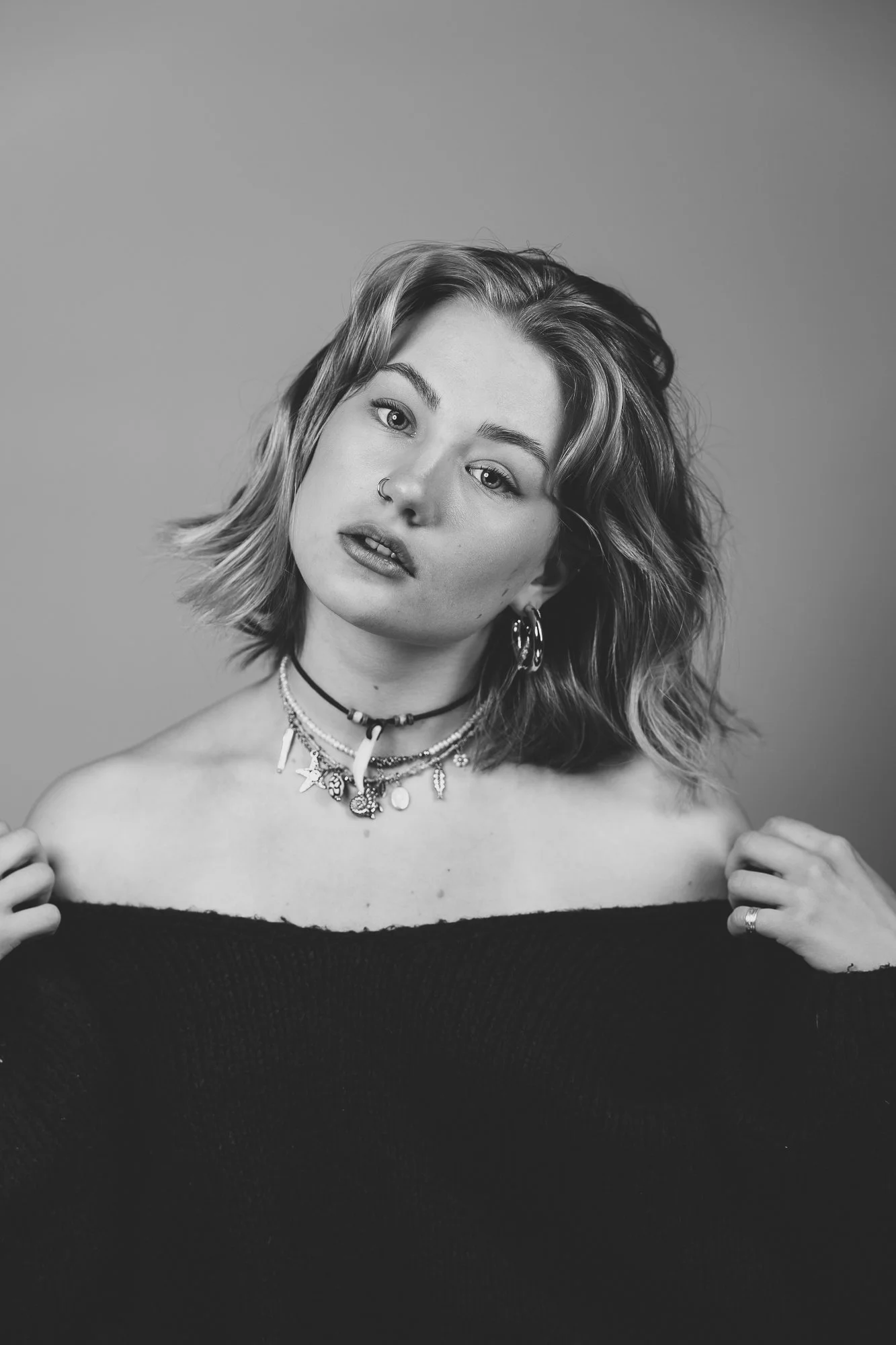 Porträt einer jungen Frau mit kurzen, gewellten Haaren, mehreren Halsketten, Ohrringen und einem Nose-Ring, die einen schwarzen Pullover trägt und den Blick direkt in die Kamera richtet.