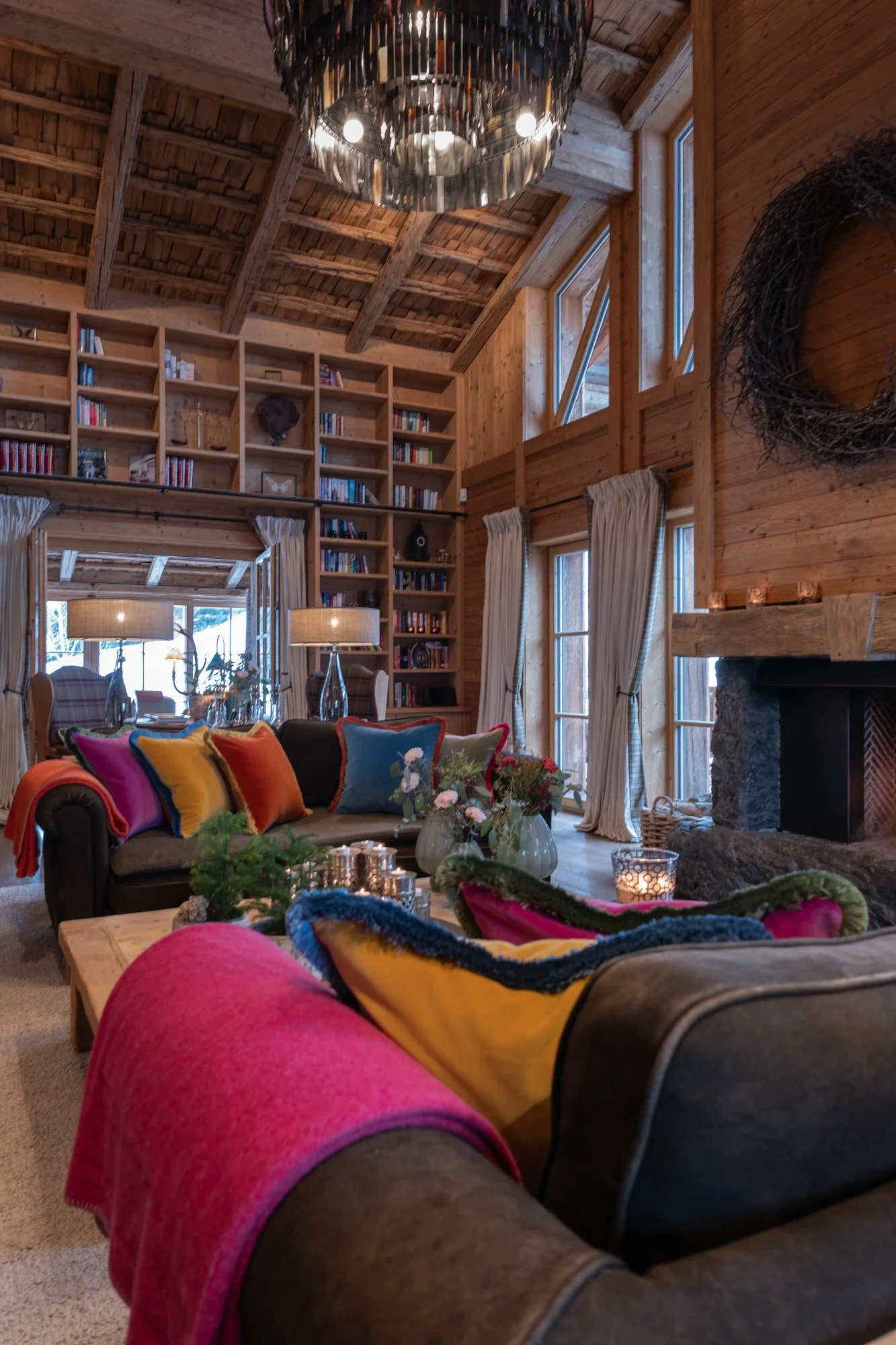 Die Schmiedalm - Premiumchalet - Gemütliches Wohnzimmer mit Holzdecke, Bücherregal, Kamin, großen Fenstern, bunten Kissen und Dekorationen.