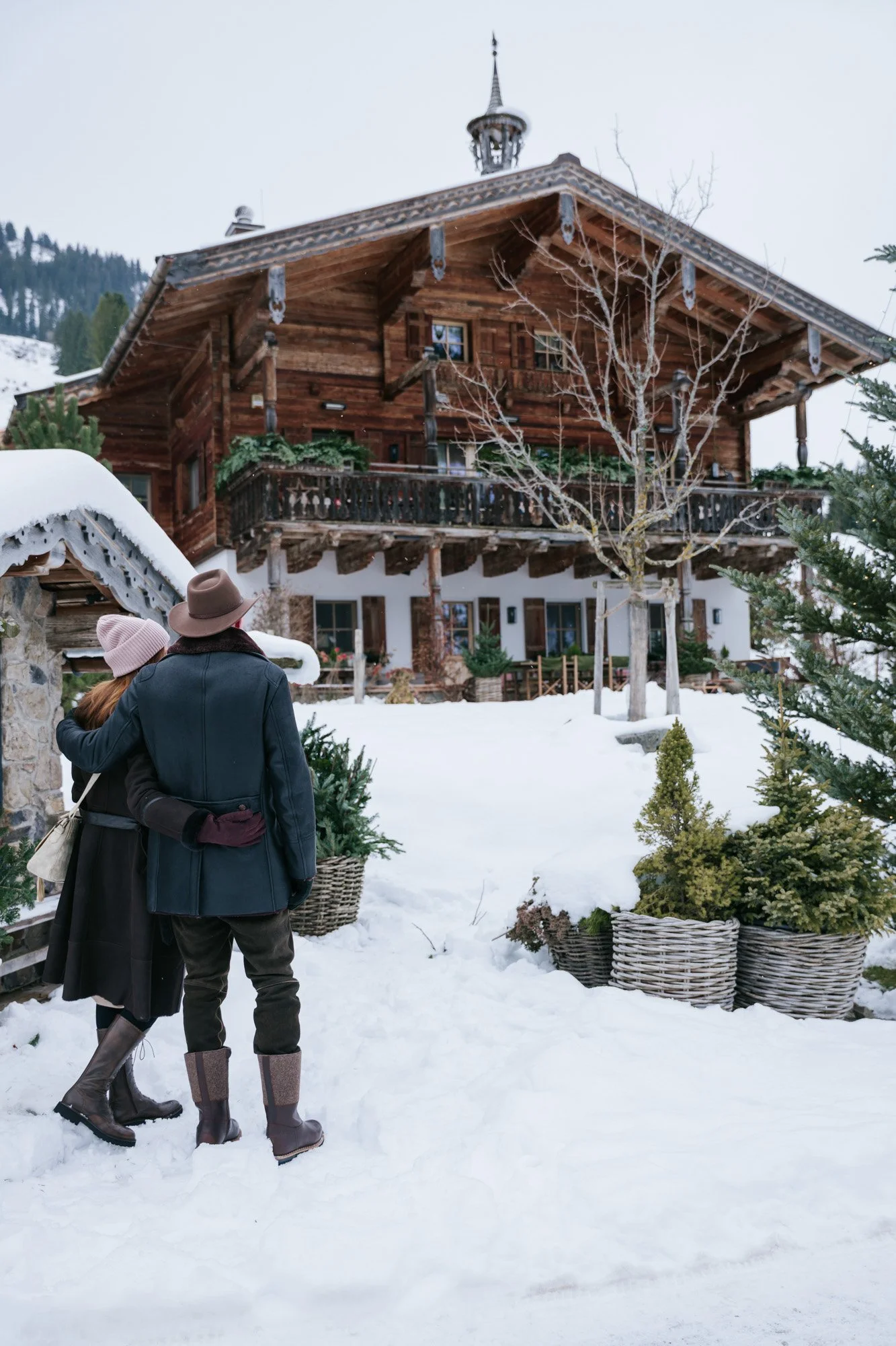 Zwei Menschen mit Winterkleidung stehen vor einem traditionellen, mehrstöckigen, holzgedeckten Chalet im Schnee.