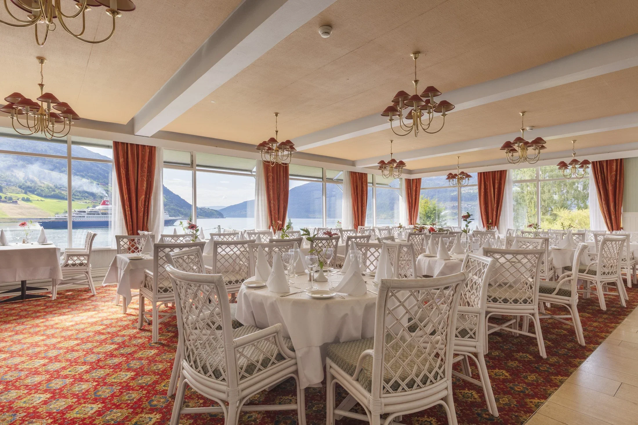 Innendørsrestaurant med store vinduer som viser fjell og vann, hvite stoler med etuiet, dekket bord med hvite duker og pyntede bord, og taklamper med flere små skærmer.