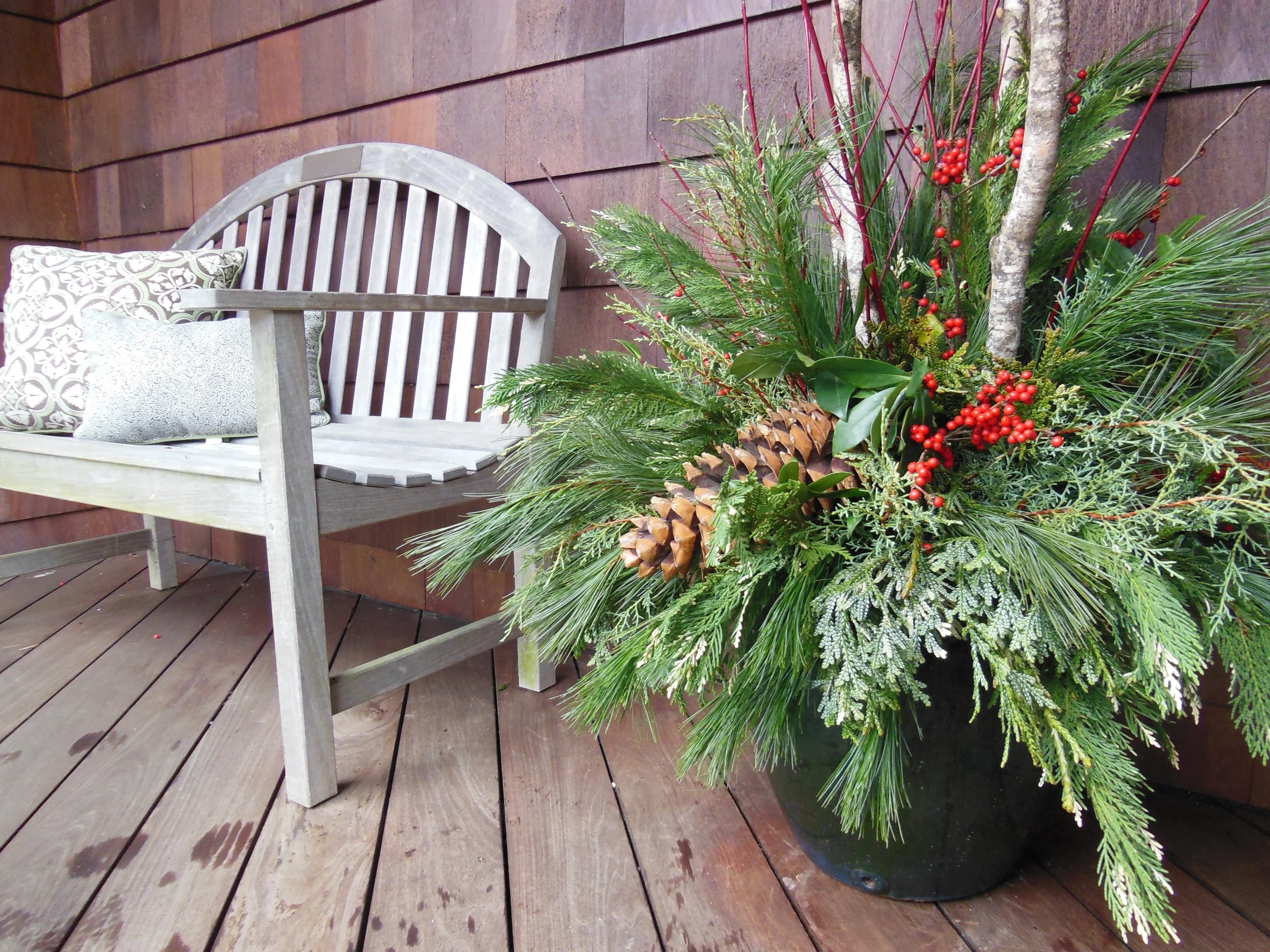 Wooden bench with patterned and plain cushions on a wooden deck, next to a large decorative planter. The planter contains pine branches, red berries, and a large pine cone, enhancing its festive appearance.