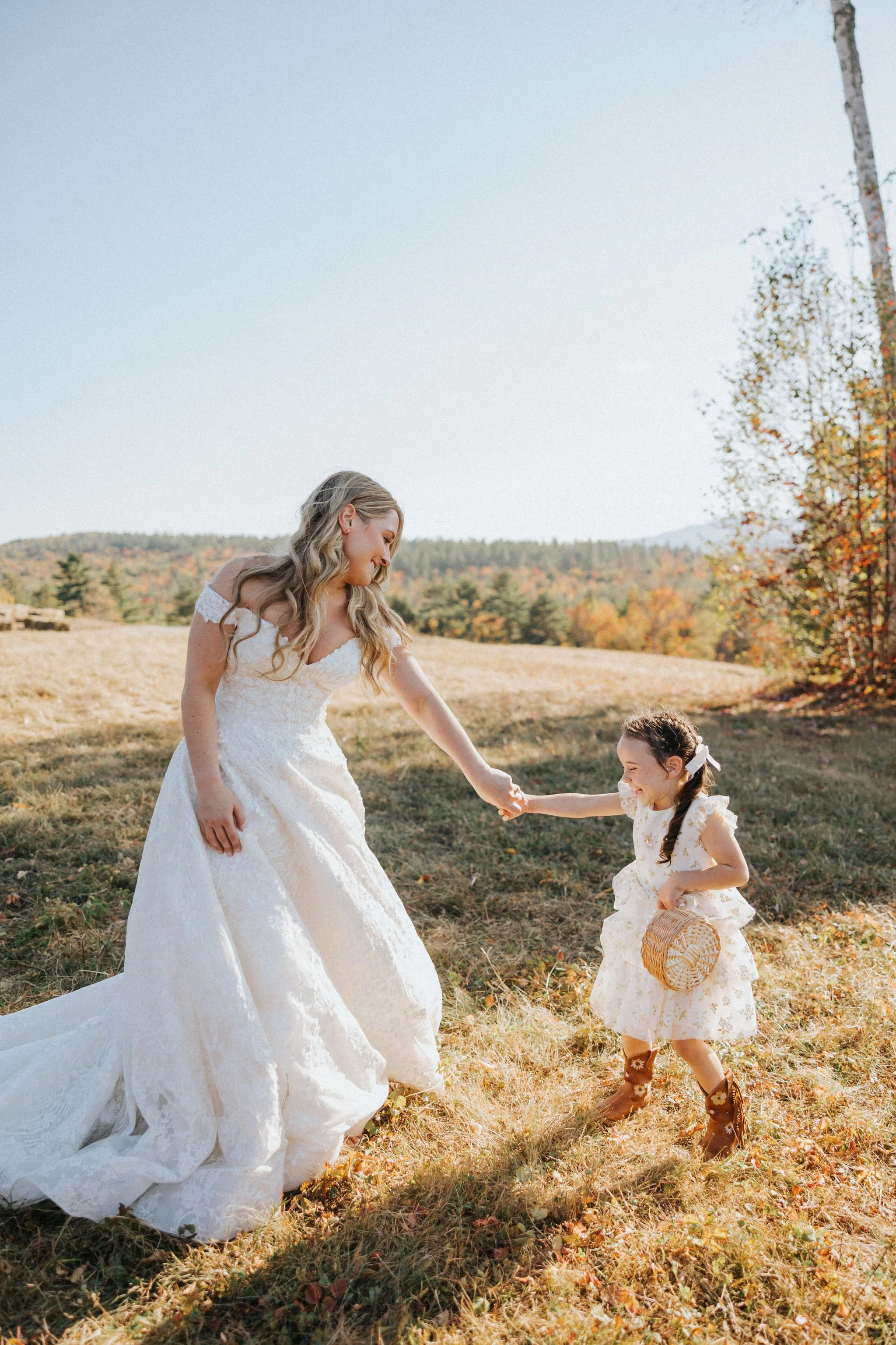 Smiling Maine bride dances with a young flower girl in a field during golden hour