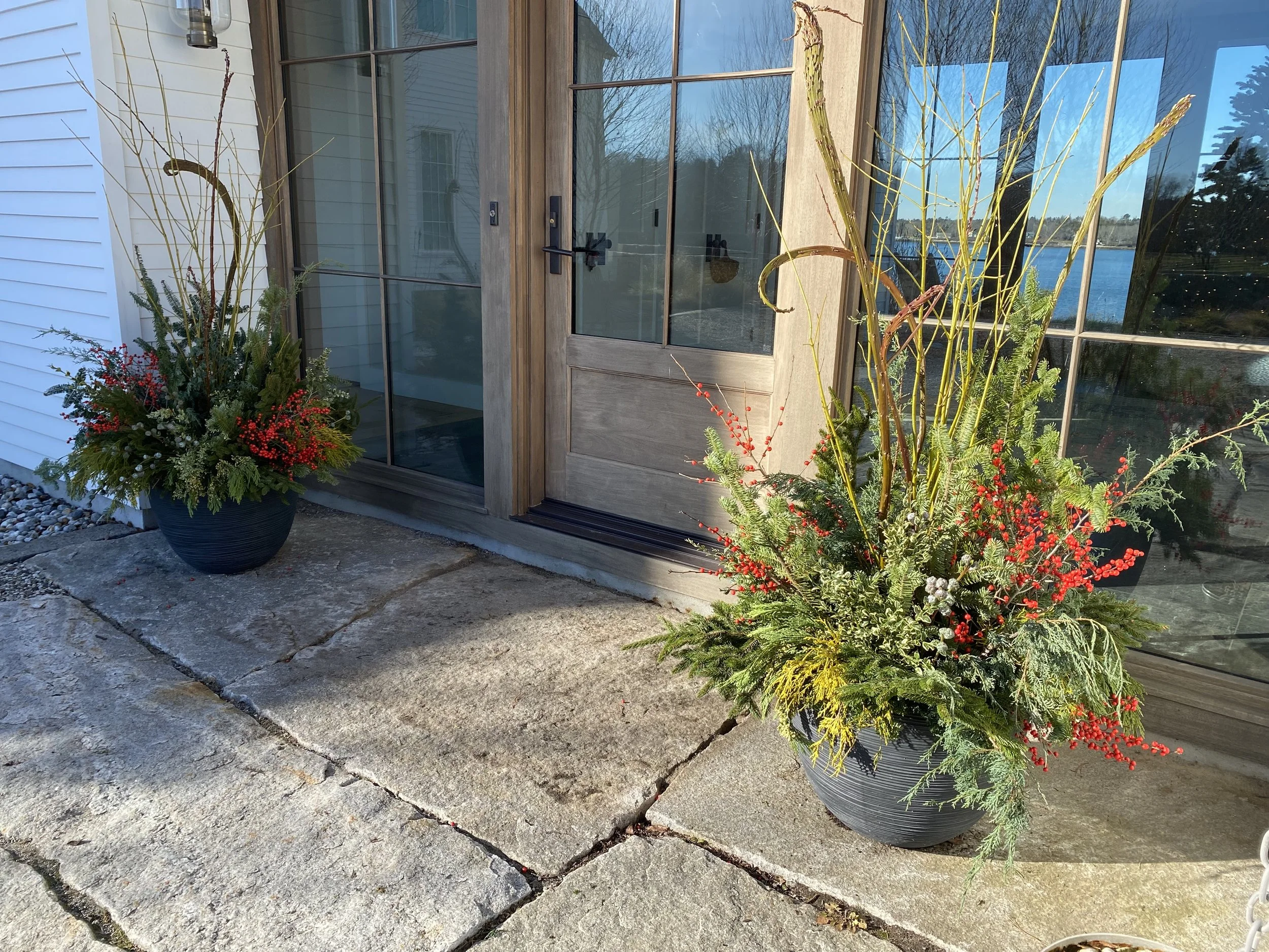 Outdoor entrance with two large planters containing winter greenery and red berries, placed on a stone patio in front of wooden double doors with glass panes.