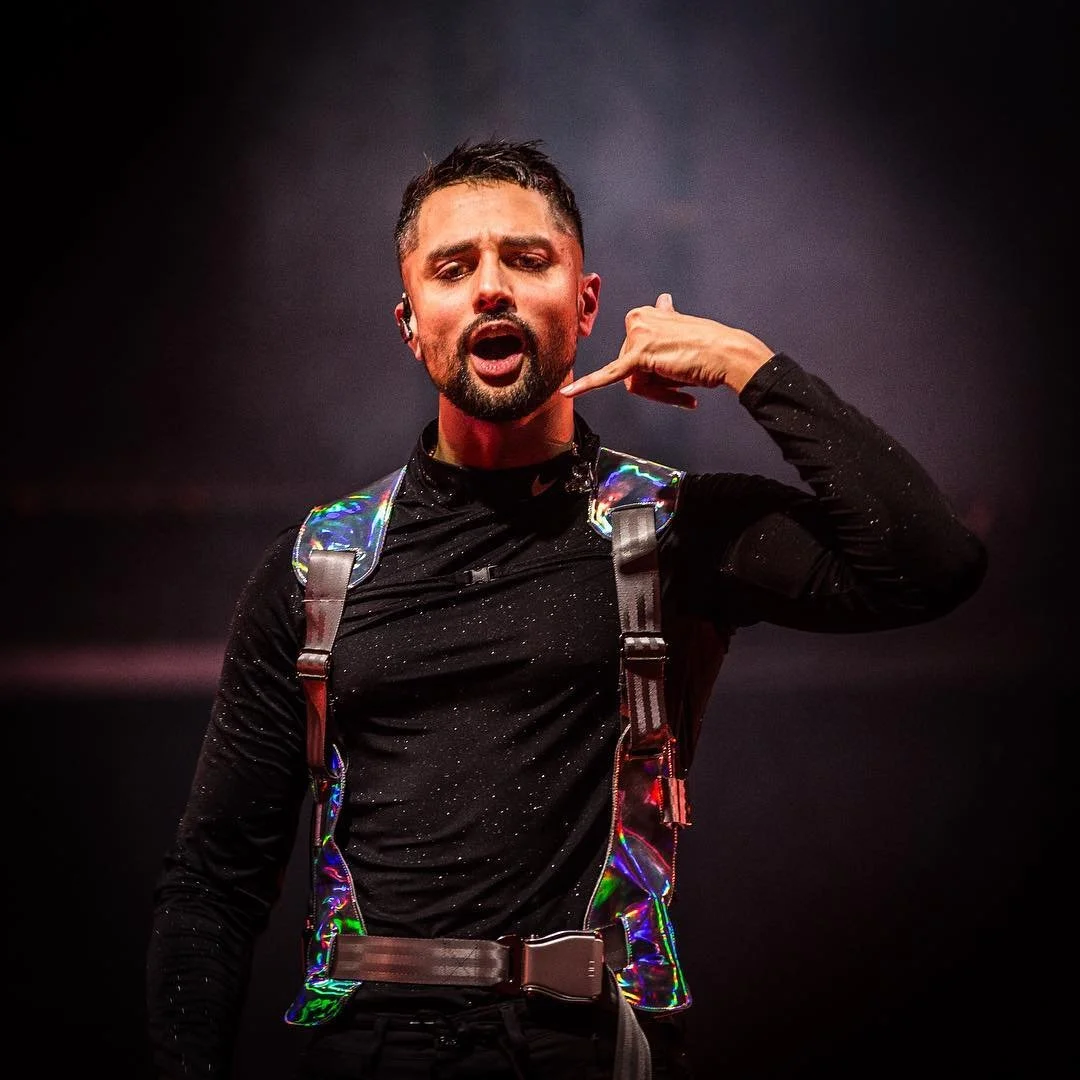 En mann som står mot en mørk bakgrunn, iført svart skjorte med glitrende detaljer.