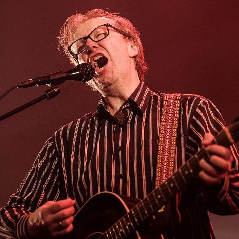 Lars Lillio Stenberg iført brun og hvit stripete skjorte og svarte briller, synger inn i en mikrofon mens han spiller gitar på scenen.