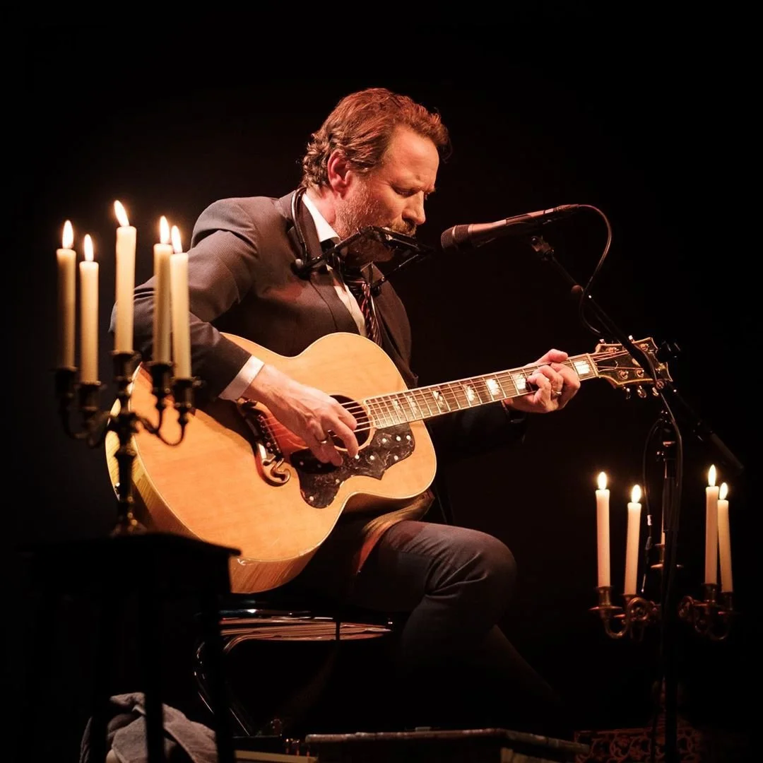 En man spiller akustisk gitar på scene med levende lys rundt.