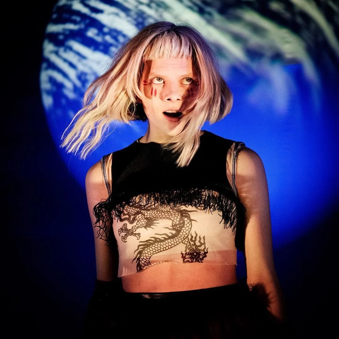 Artisten Aurora med blondt hår og tatovering av en drage på overkroppen, som står foran en blå bakgrunn.