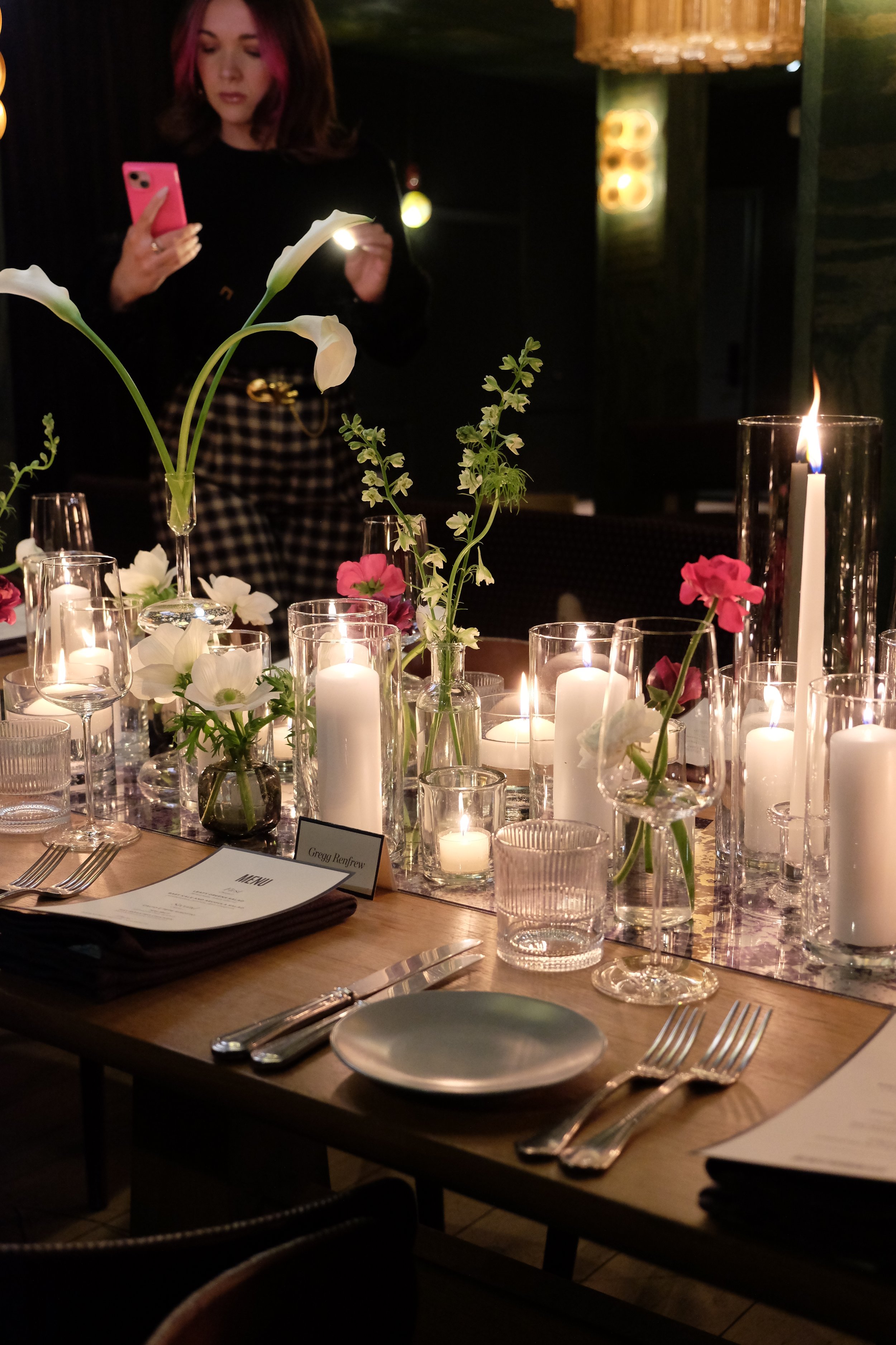 A dinner table decorated with white and pink flowers in glass vases, surrounded by lit candles in various glass holders, with place settings including forks, knives, a plate, and menus. A woman in black top and checkered pants is in the background, l