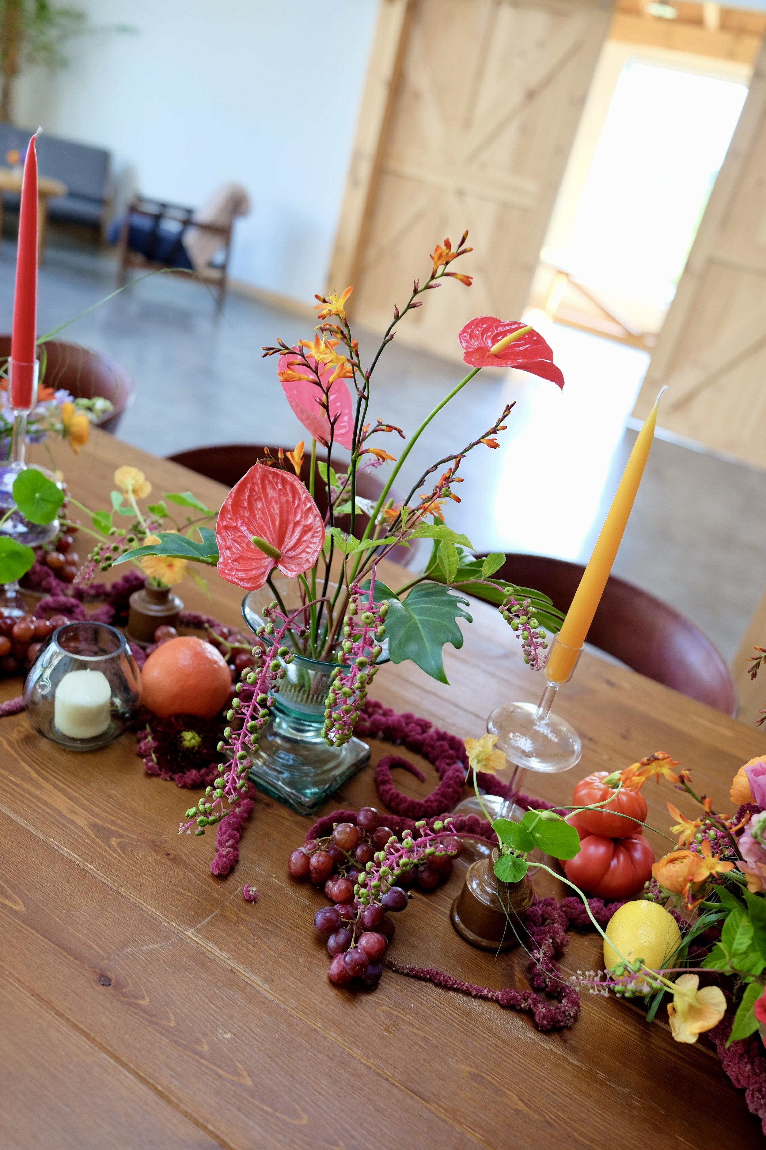 A table arrangement featuring a floral centerpiece with pink, red, and orange flowers, surrounded by grapes, apples, and candles, situated on a wooden table with a rustic interior background.
