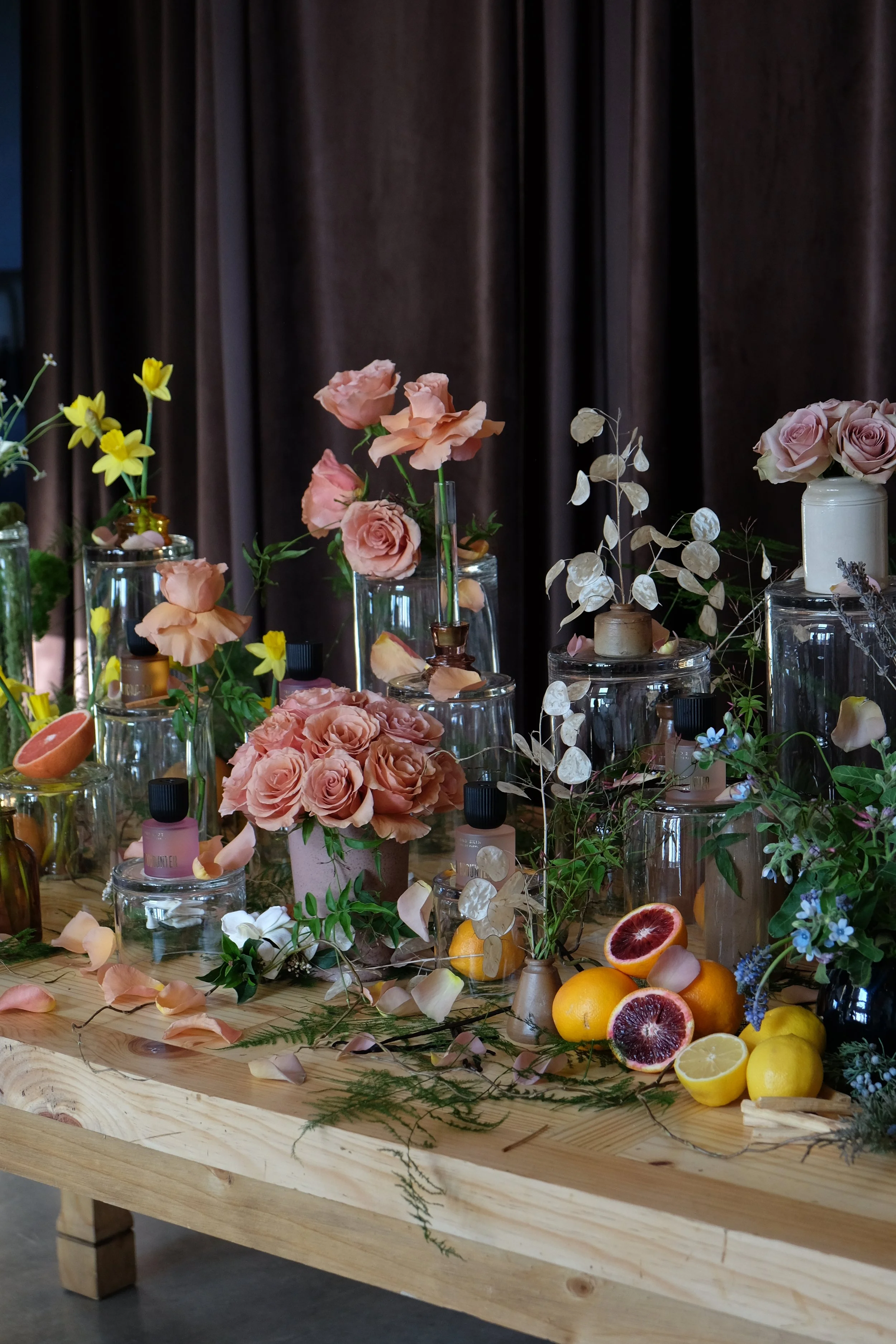 Floral arrangement with pink roses, yellow flowers, and citrus fruits on a wooden table with dark curtains in the background.