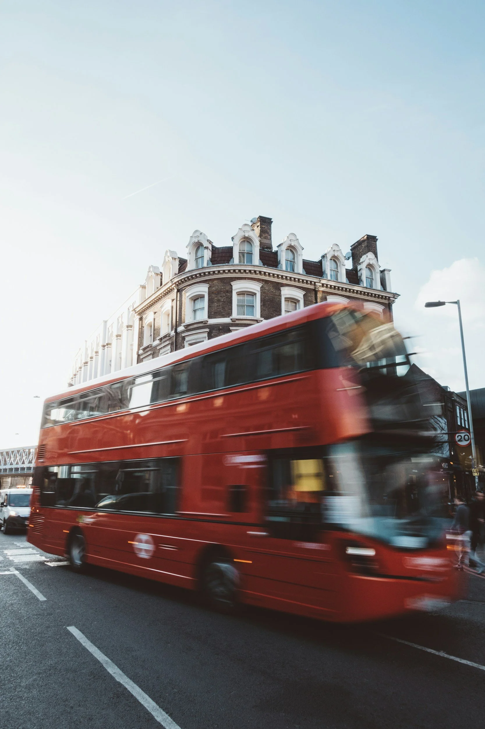 A red double-decker bus moving fast on a city street in front of a tall, historic building with a curved facade and multiple windows. The sky is clear and blue, and there is a streetlamp visible on the right side.