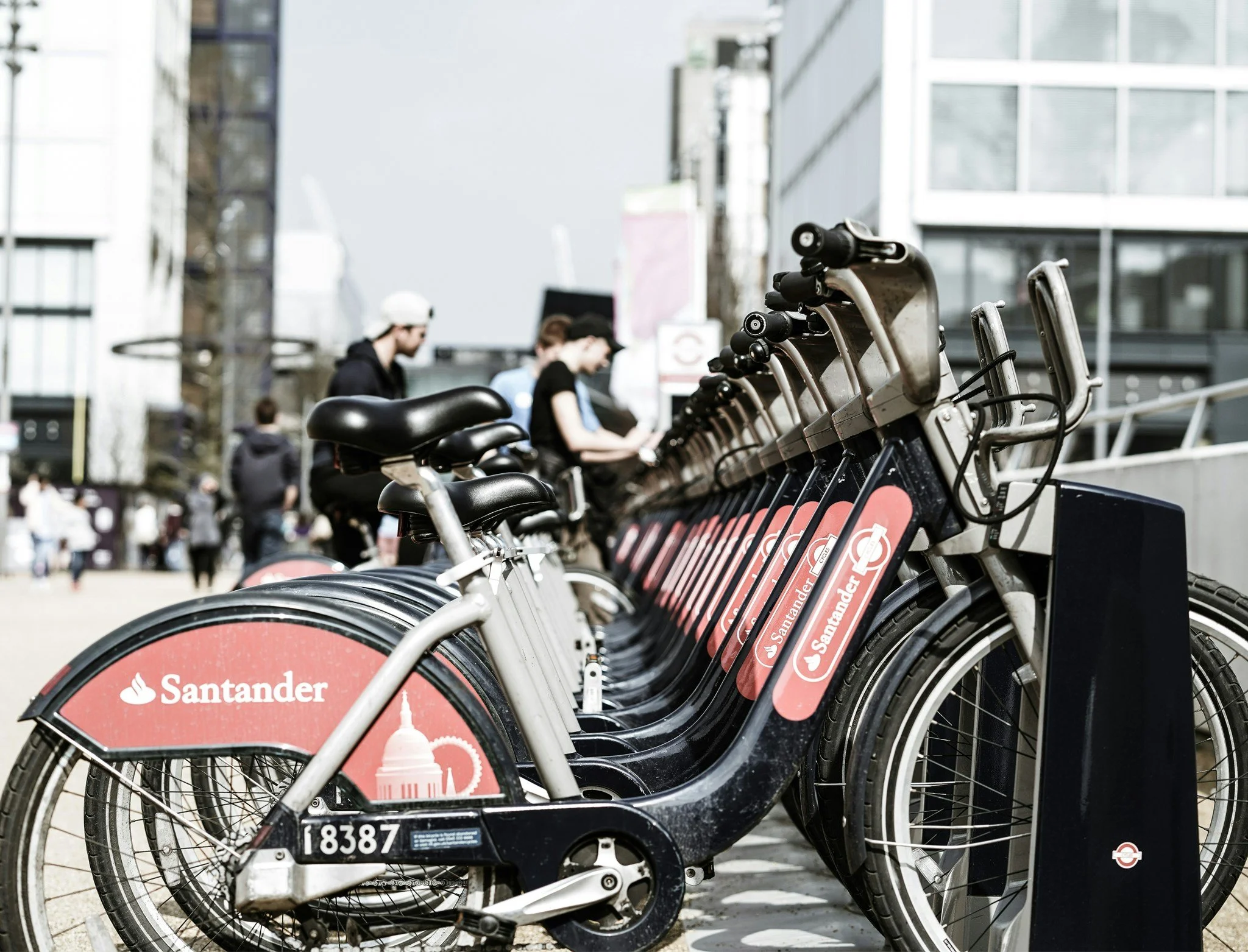 Bike-sharing bikes with Santander branding parked in a row on a city sidewalk, with buildings and people in the background.