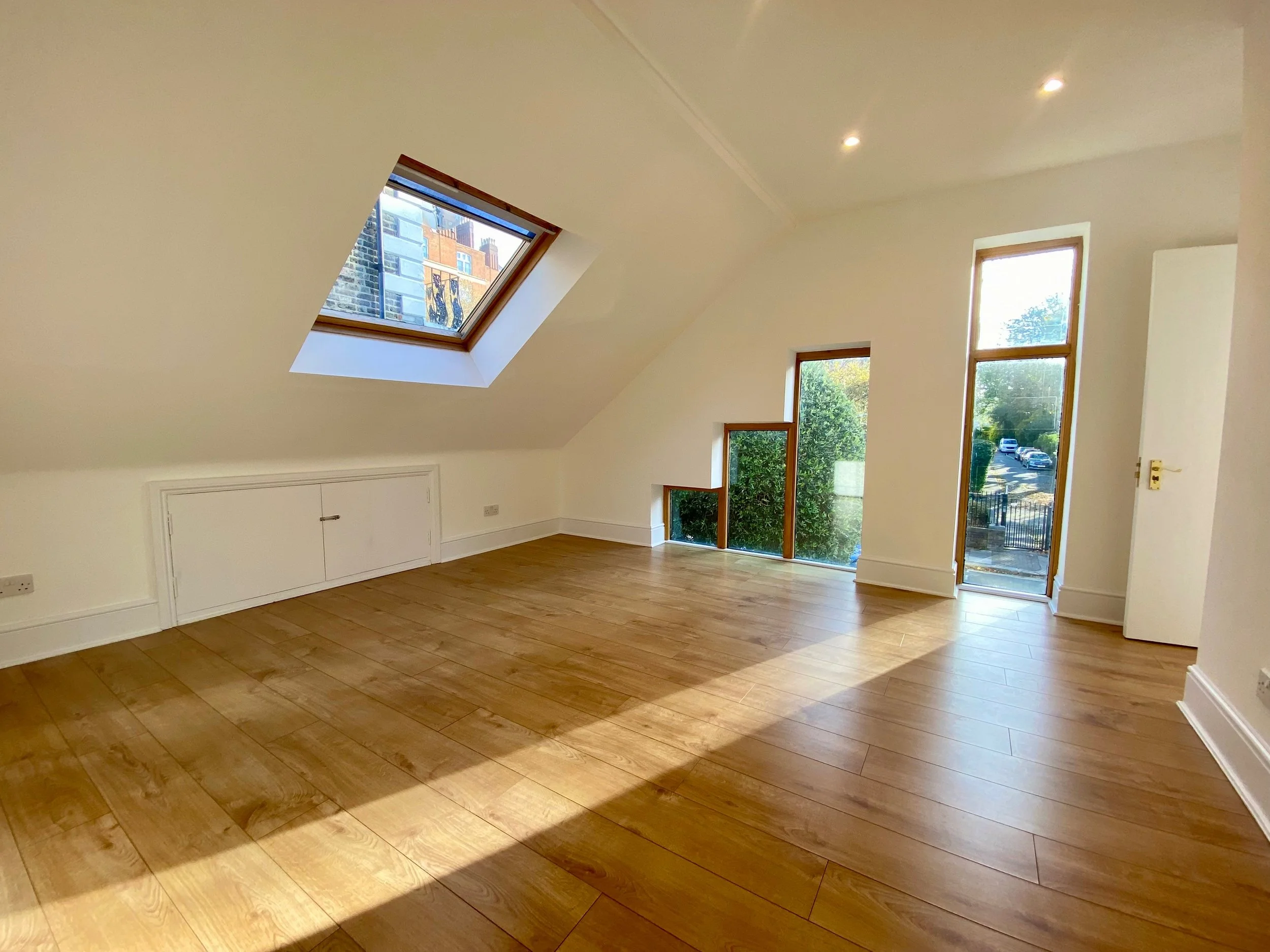 Empty room with wooden flooring, white walls, a skylight window on the sloped ceiling showing external brick buildings, and large windows letting in sunlight and showing greenery outside.