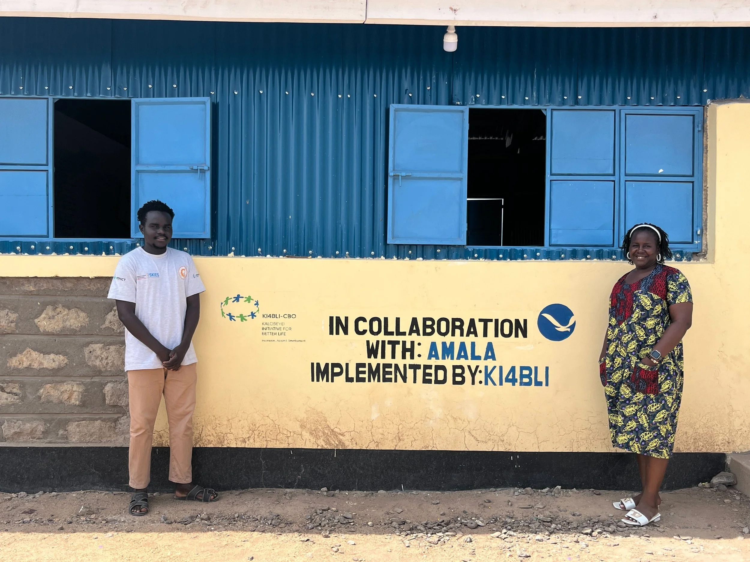 Two people standing either side of a wall that says 'In collaboration with Amala Implemented by: Ki4Bli'