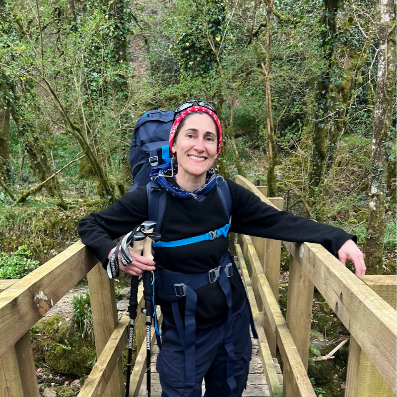 A smiling woman with a backpack standing on a wooden bridge in a wooded area.