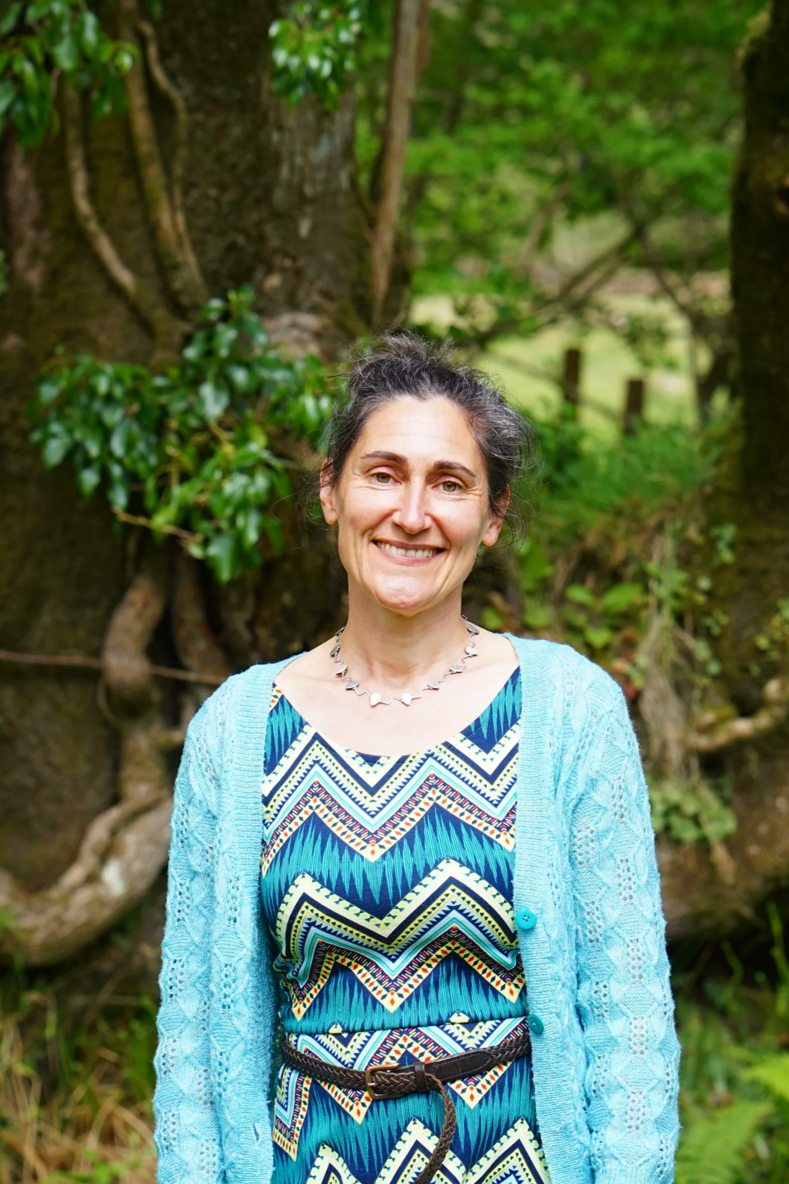 A smiling woman with a backpack standing on a wooden bridge in a wooded area.