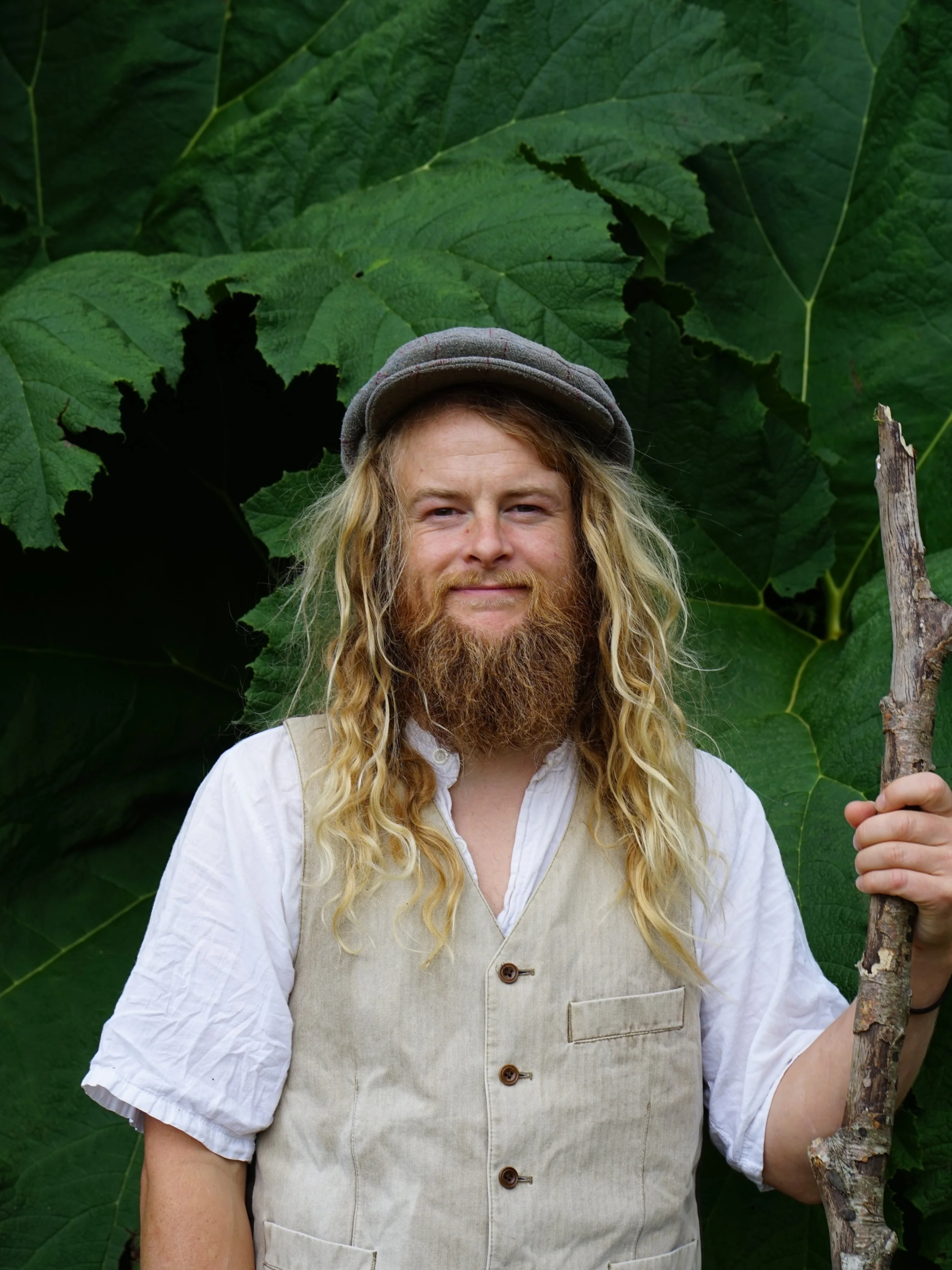 A man with long blonde hair, a full beard, wearing a gray cap, a blue shirt, and a dark vest standing in front of a wooden wall.