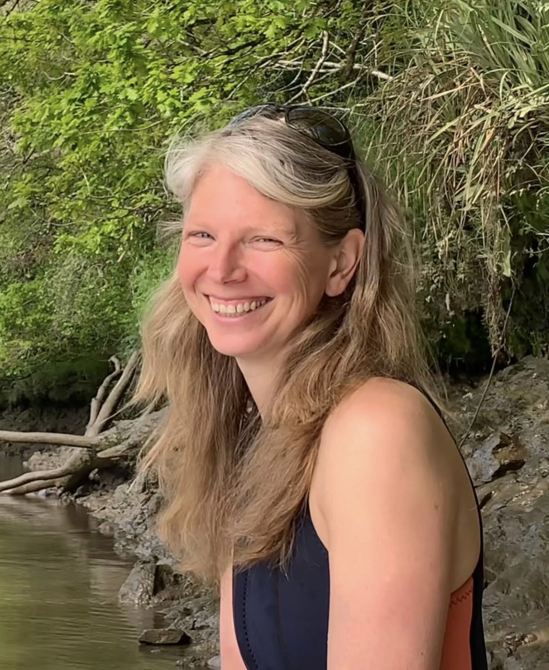 A young woman standing outdoors beside a wheelbarrow filled with two young children, one girl and one boy, in a garden area with a stone wall and window behind. The boy has a pacifier and is dressed in a green raincoat, while the girl has blonde hair and is wearing a pink coat.