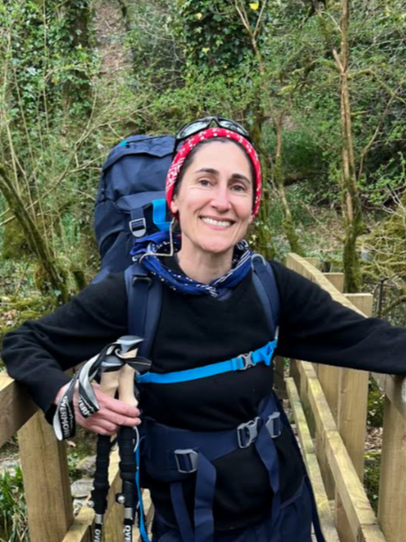 A smiling woman with a backpack standing on a wooden bridge in a wooded area.