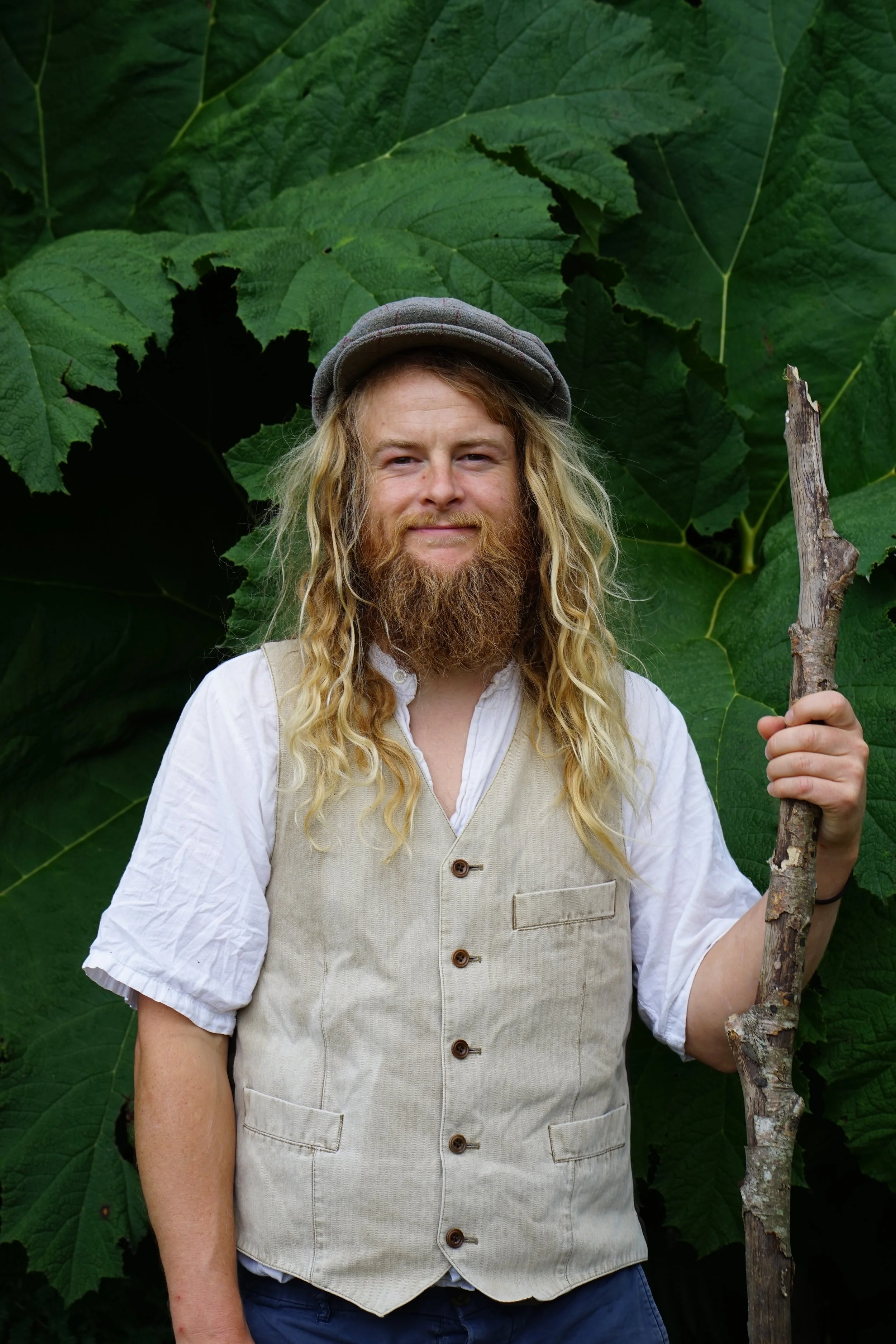 A man with long blonde hair, a full beard, wearing a gray cap, a blue shirt, and a dark vest standing in front of a wooden wall.