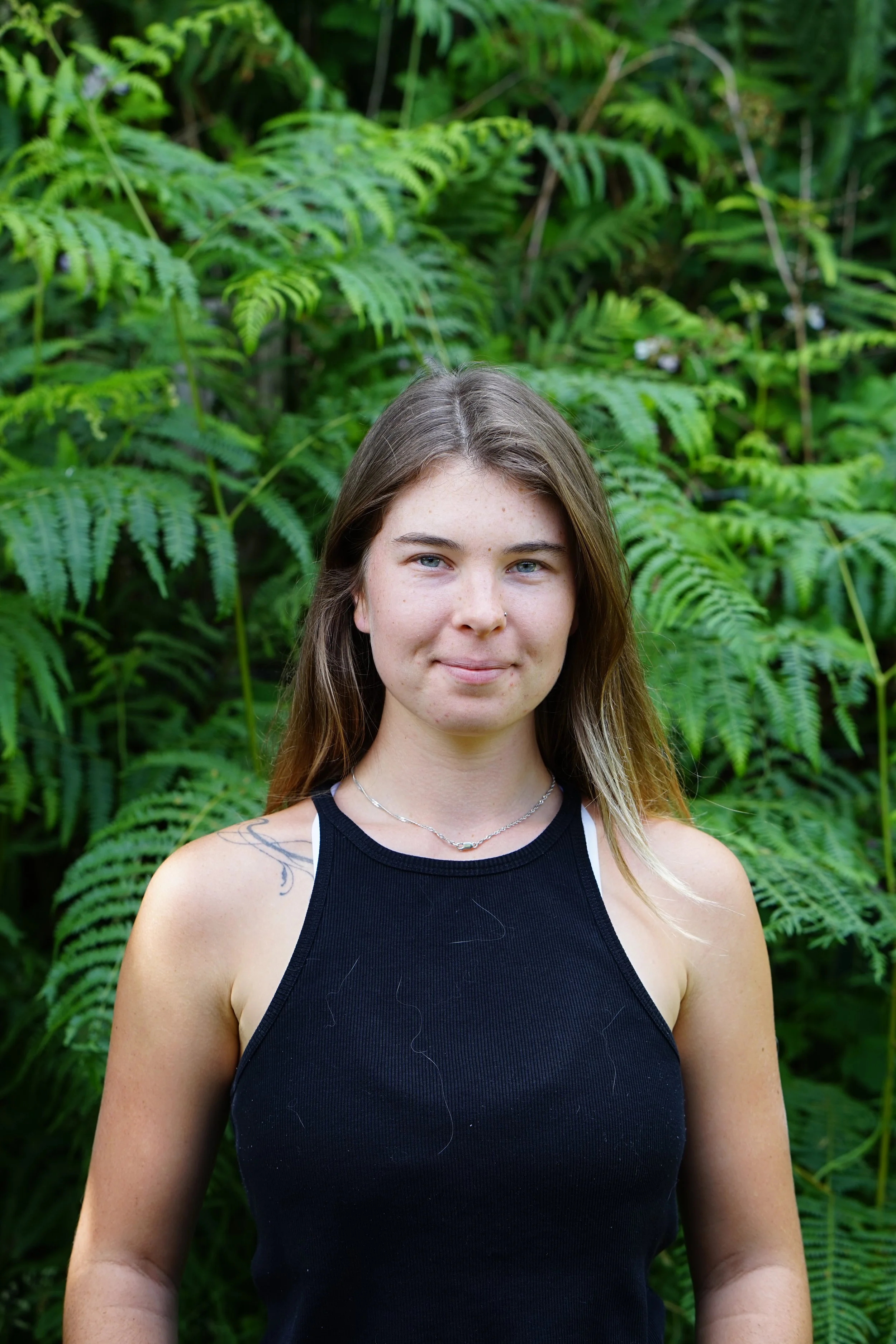 A young woman with long blonde hair and a white t-shirt standing outdoors in front of a large tree with a wooden fence and green plants in the background.