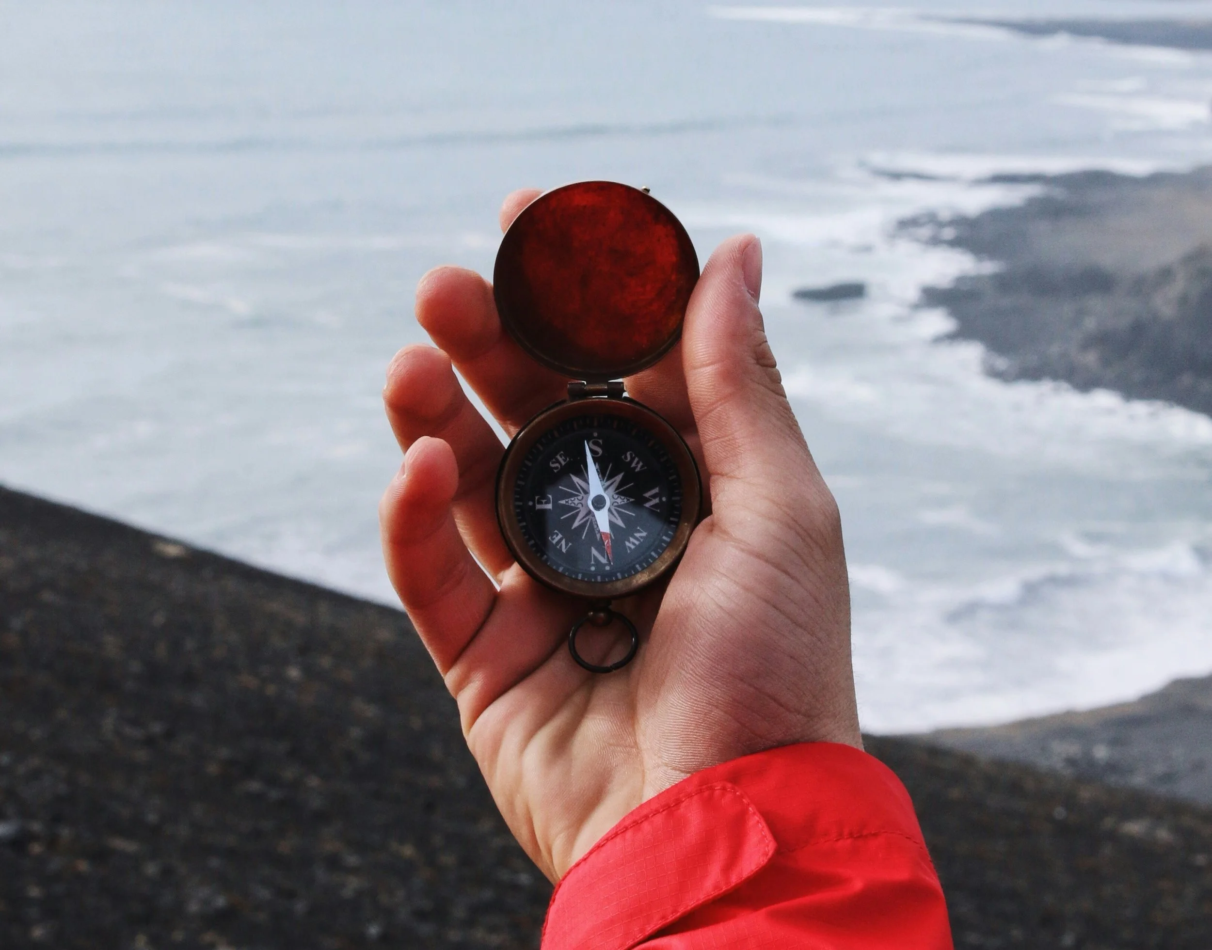 A hand holds a compass, pointing South, with a coastal shoreline in the background