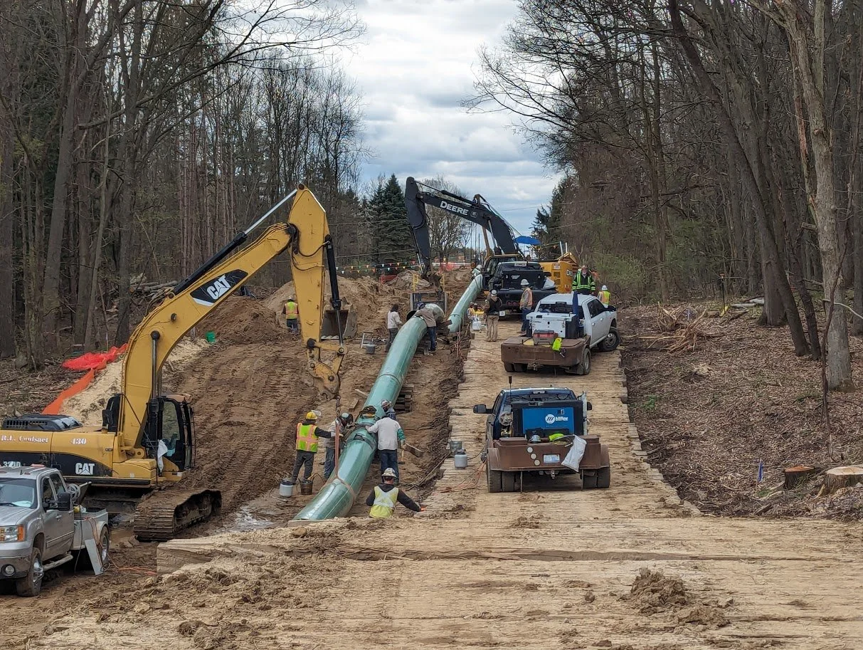R.L. Coolsaet workers installing large pipeline section in wooded area with excavators.