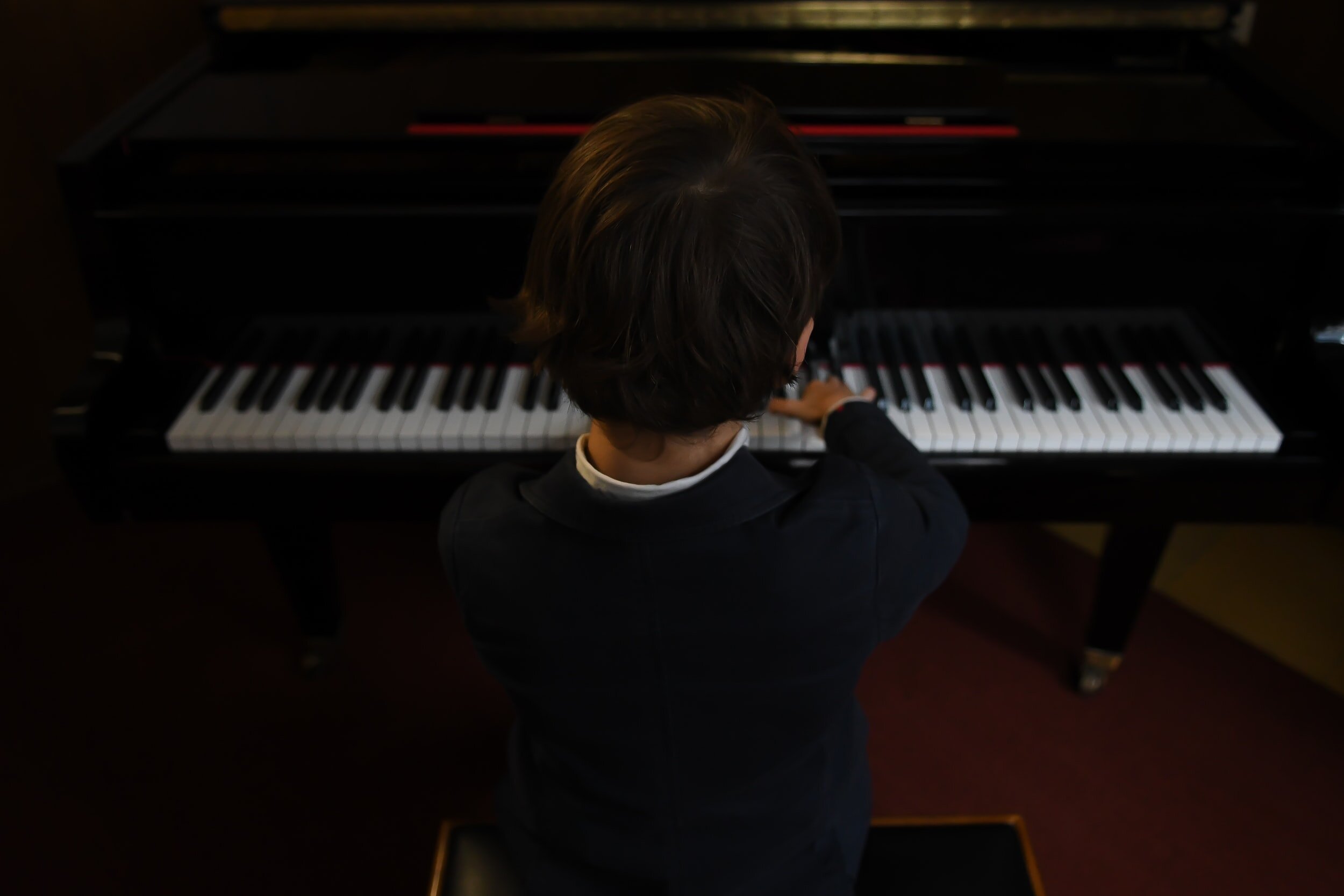 A person with dark hair wearing a dark suit jacket playing a black upright piano, viewed from above.