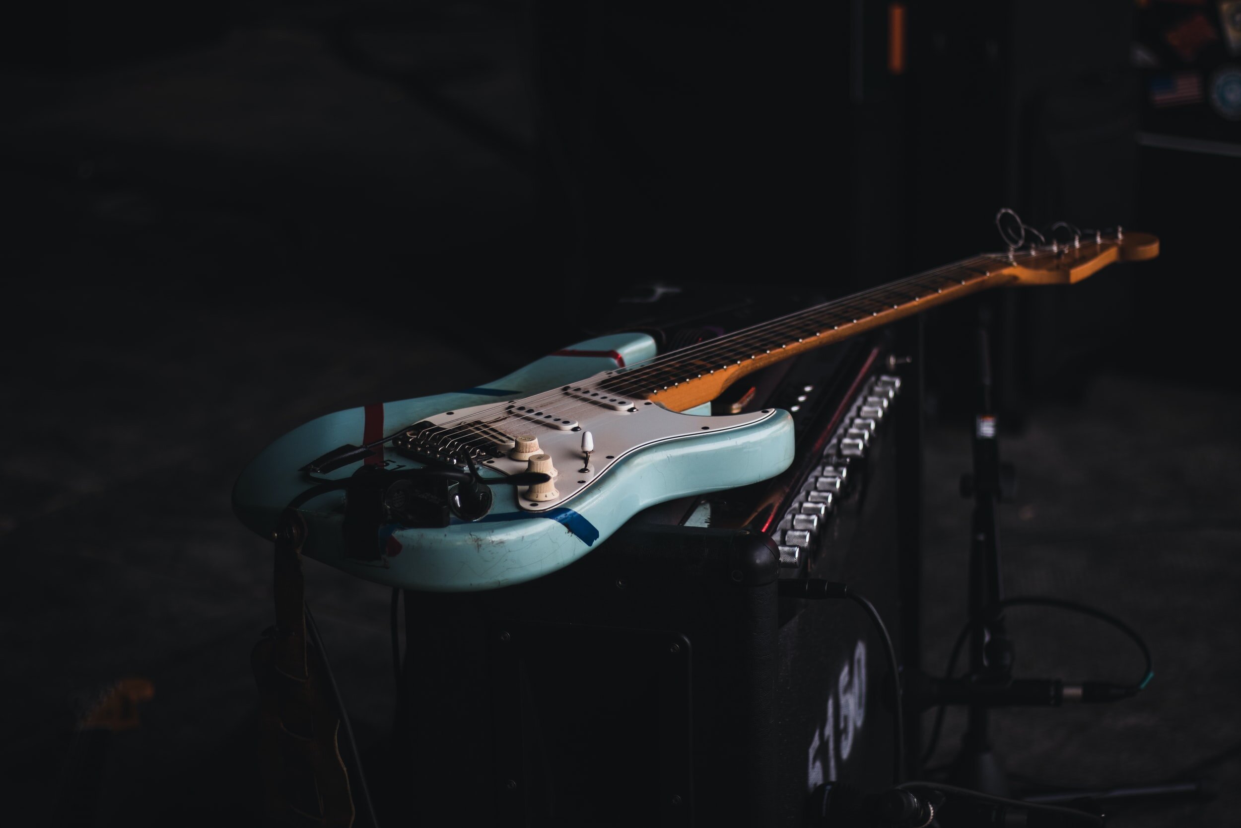 An electric guitar with a turquoise body, cream-colored pickguard, and maple neck rests on a stand in a dark room, with a keyboard partially visible underneath.