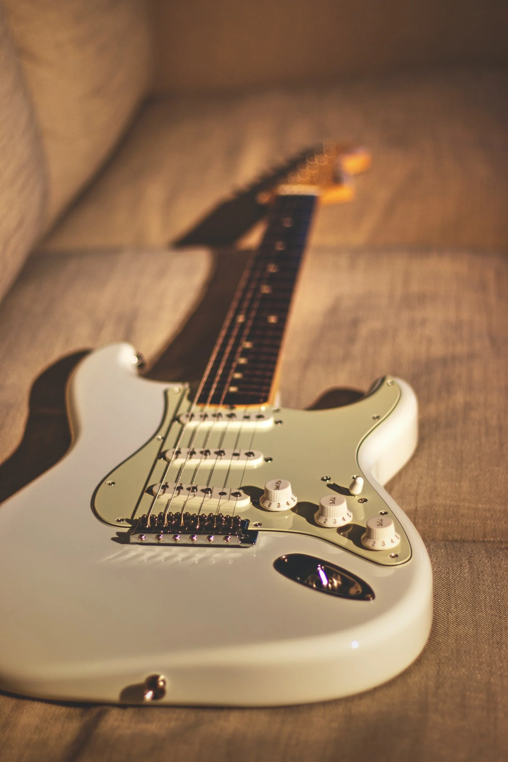 A close-up of a white electric guitar resting on a beige fabric surface, with a blurred background.