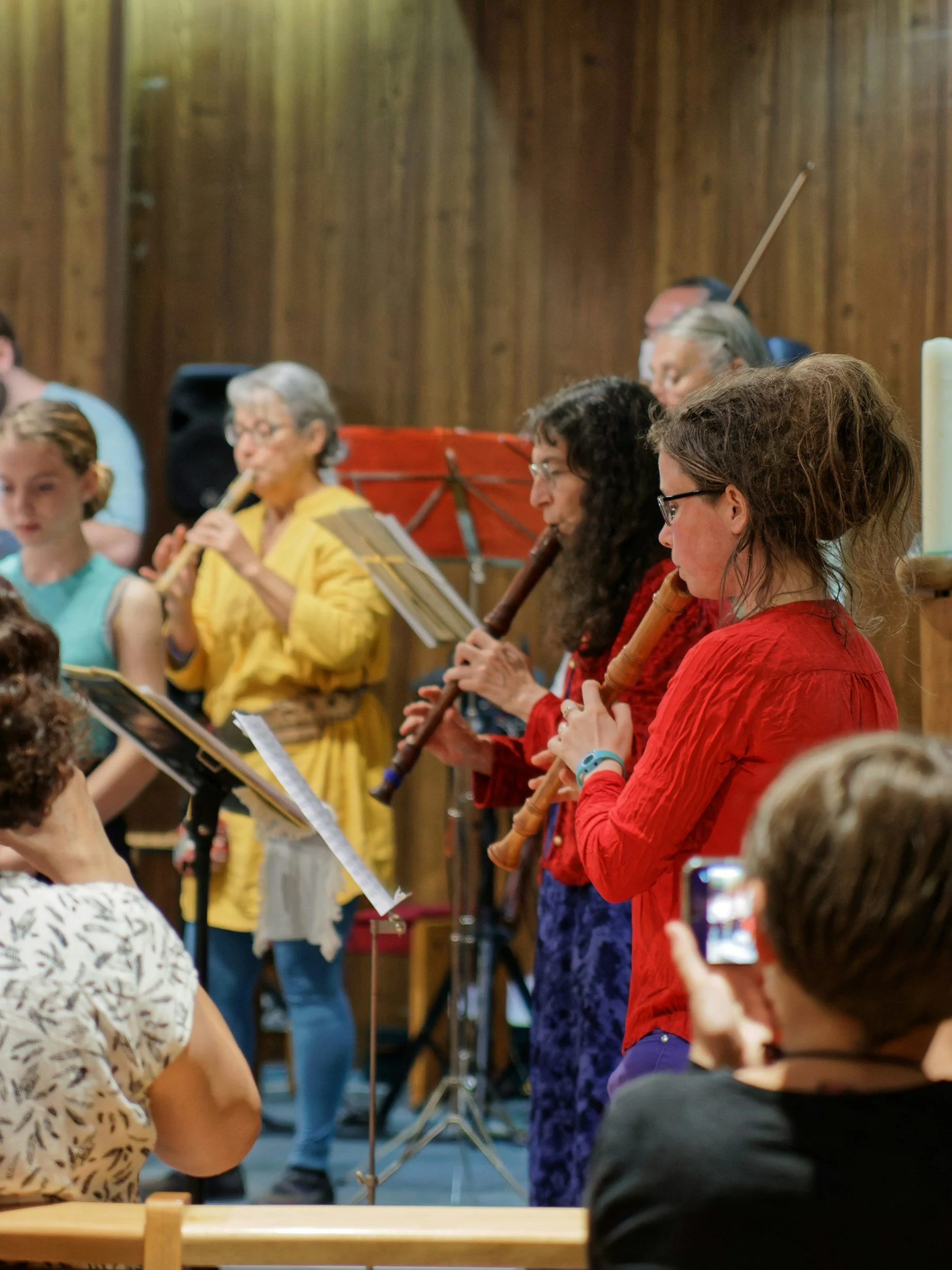 Group of people, predominantly women, playing recorders during a performance in a wood-paneled room.