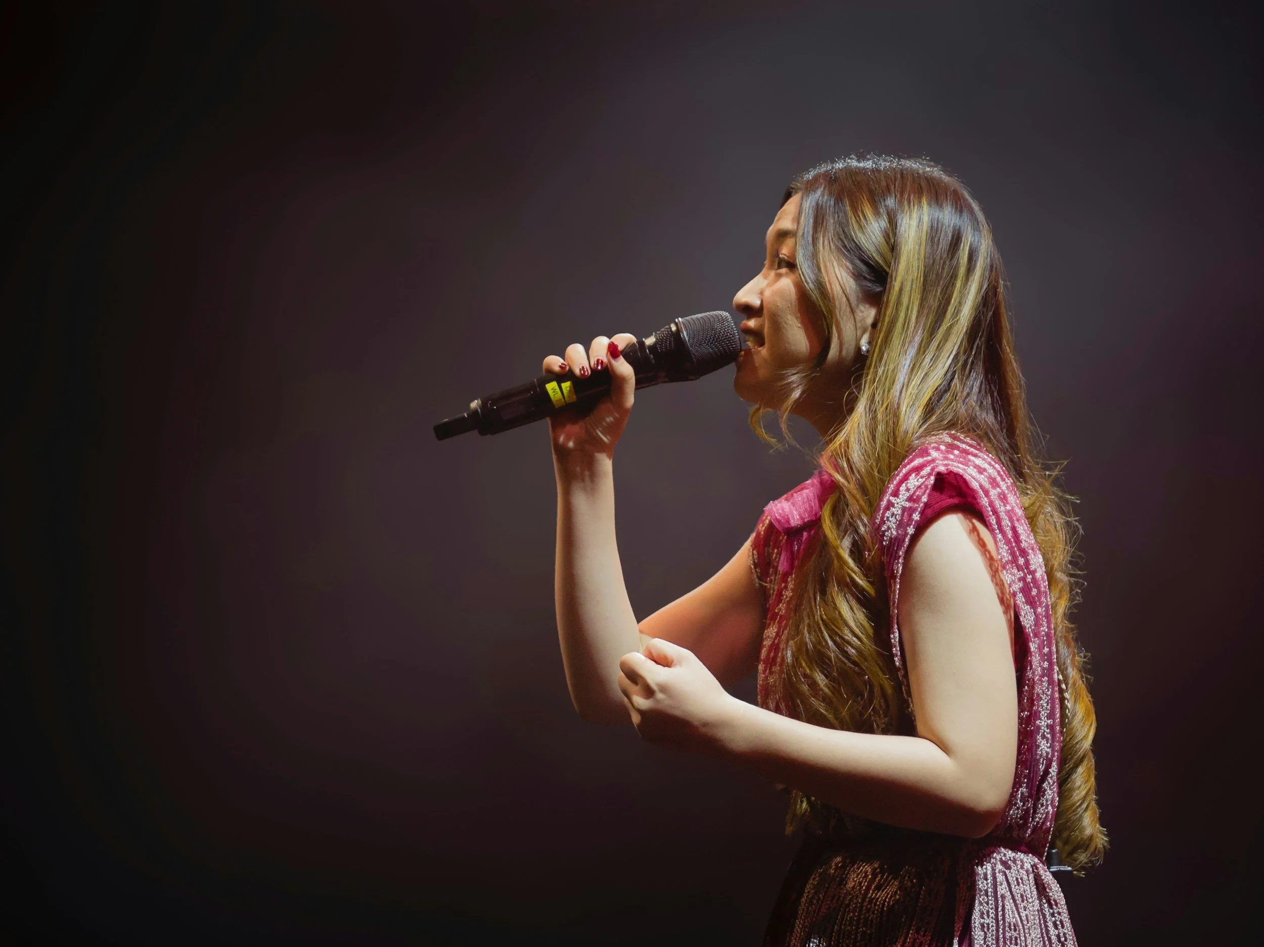 A woman with long wavy hair and earrings singing into a black microphone on stage, dressed in a pink lace dress, against a dark background.
