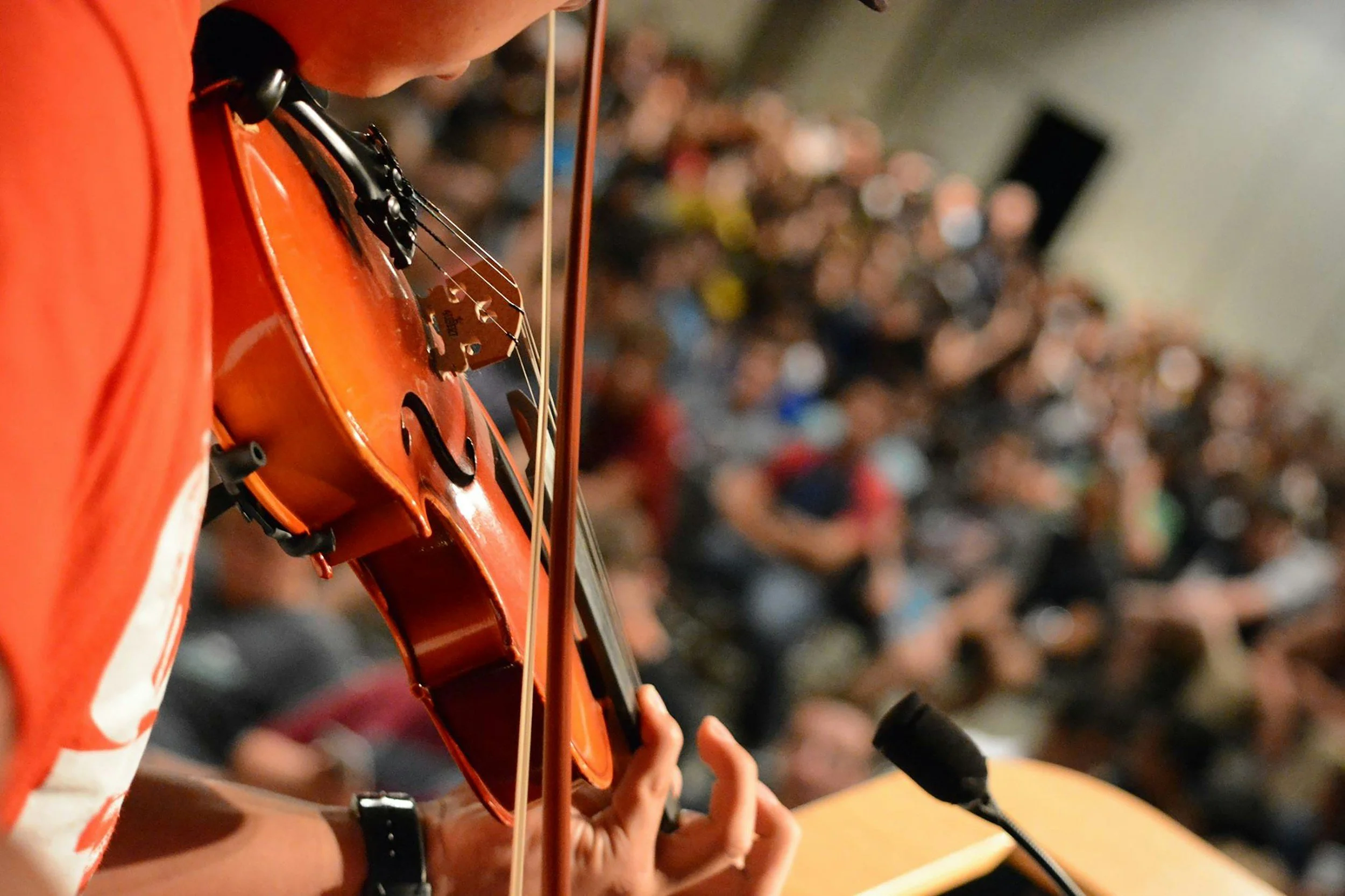 Person playing a violin, wearing orange clothing, with a large crowd blurred in the background.