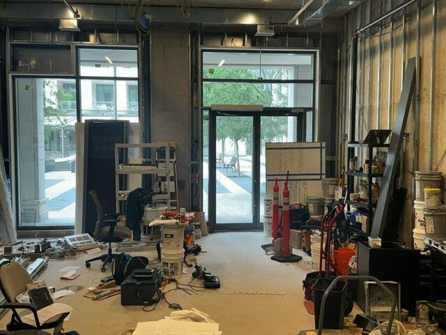 Construction site with open doorway, scattered tools and equipment, unfinished walls, and windows looking outside onto a courtyard.