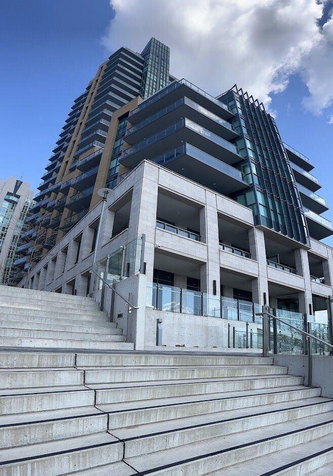 Modern high-rise building with glass balconies and wide stone steps, viewed from below under a blue sky with clouds.