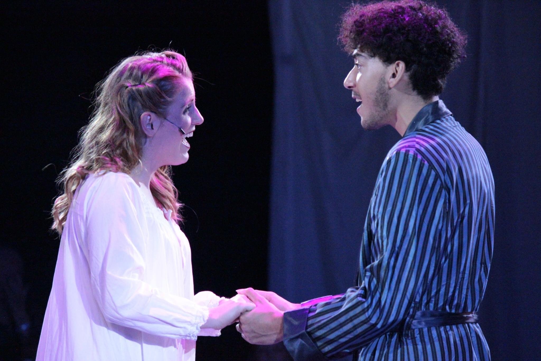 Two actors on stage holding hands, smiling and looking at each other during a theatrical performance. The woman has red hair and is wearing a white dress, and the man has curly hair and is wearing a striped jacket.