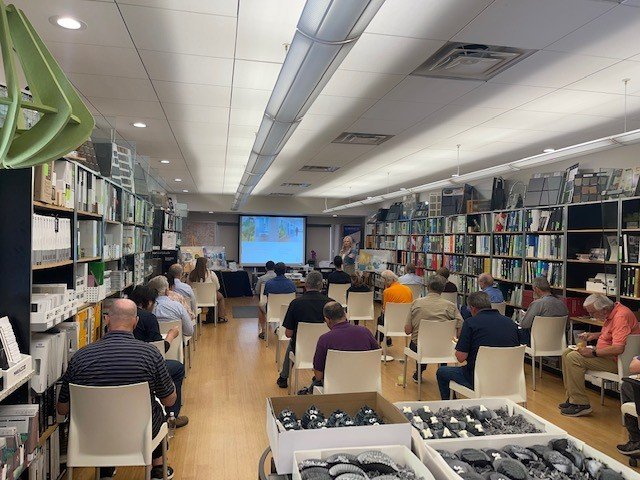 People attending a presentation at The Reference Library, seated in rows facing a large screen at the front, surrounded by bookshelves.