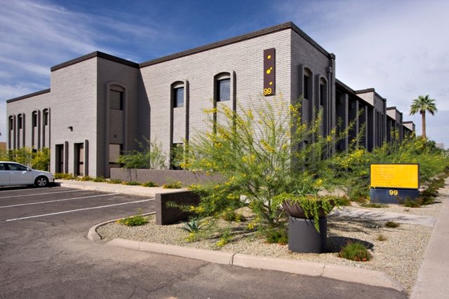 A two-story office building with a parking lot and desert landscaping in front, including a young tree and a large yellow sign.