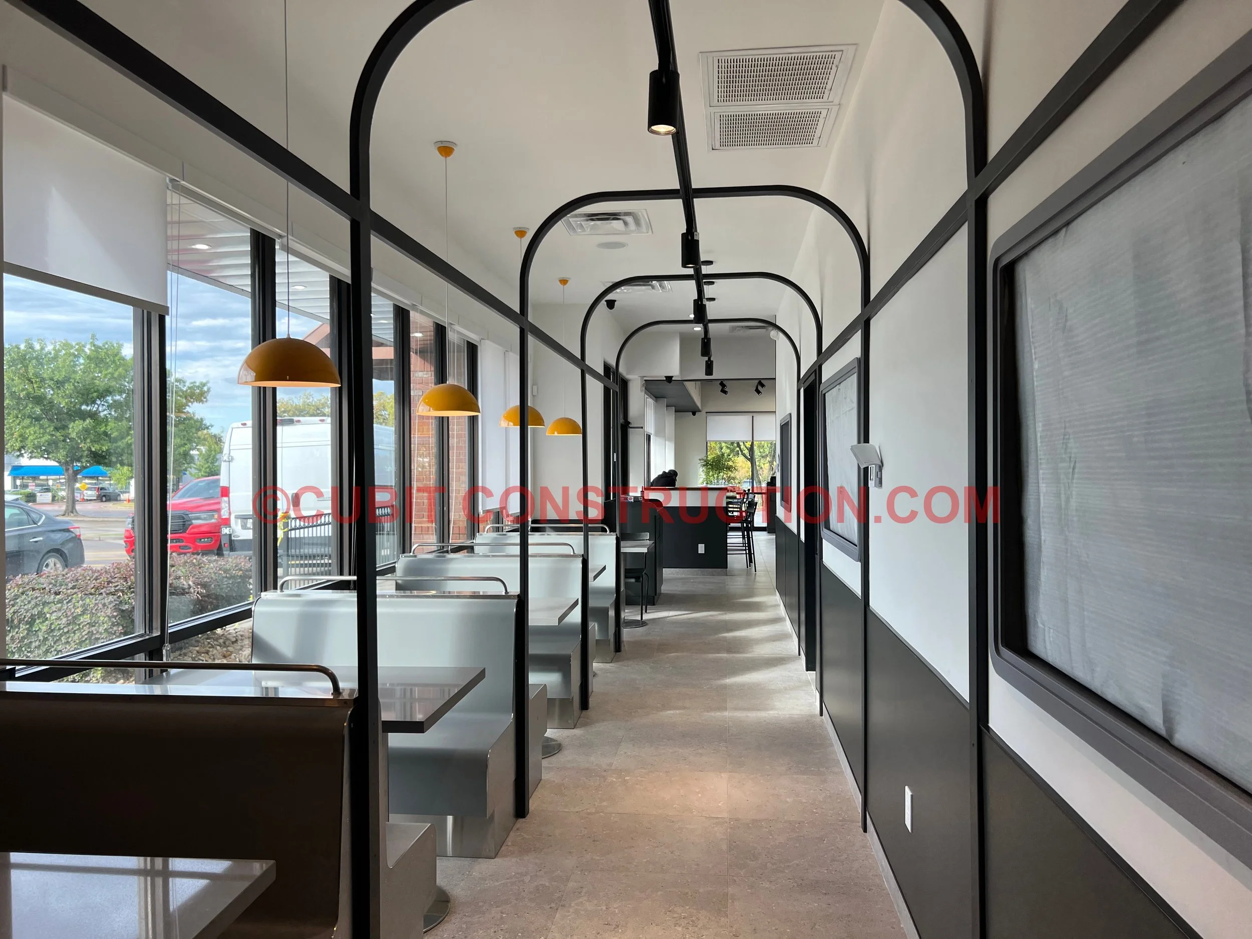 Empty modern fast-food restaurant with white booths, black railings, and yellow pendant lights, large windows showing parked cars outside.