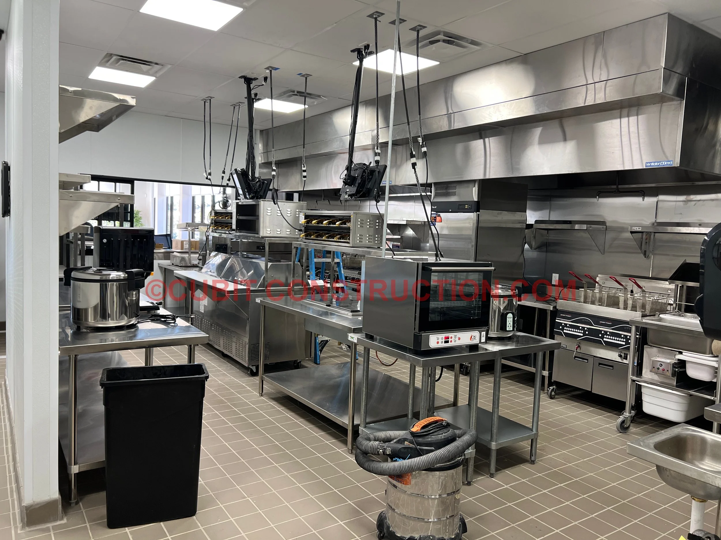 Commercial kitchen with stainless steel appliances, countertops, and equipment, including a stove, microwave, and fryers, with a vacuum cleaner in the foreground and large windows letting in natural light.