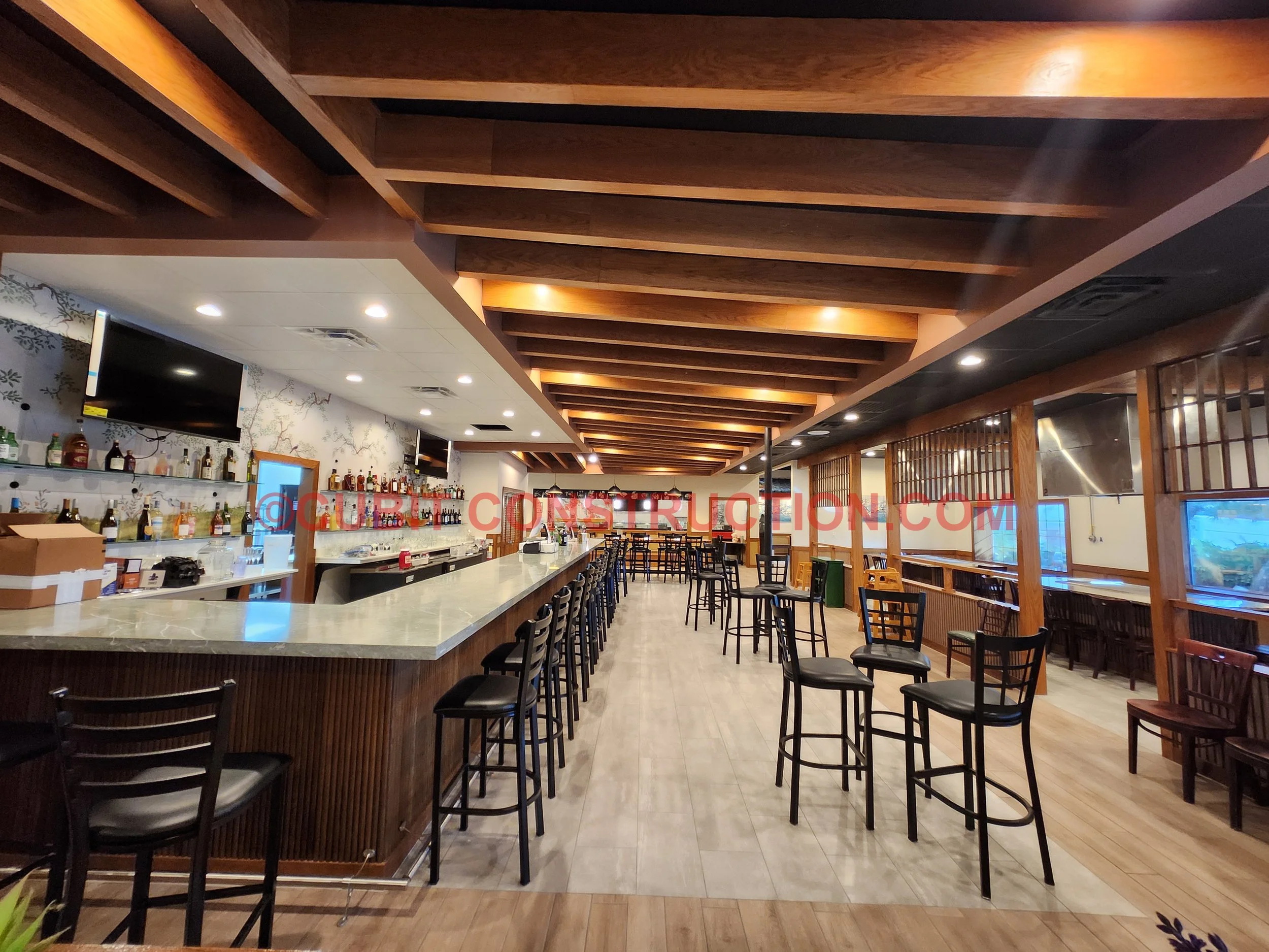 Interior of a restaurant bar with a long marble countertop, bar stools, wooden ceiling beams, and multiple tables and chairs. TV screens are mounted on the wall, and shelves behind the bar display bottles of alcohol. Large windows provide natural light.