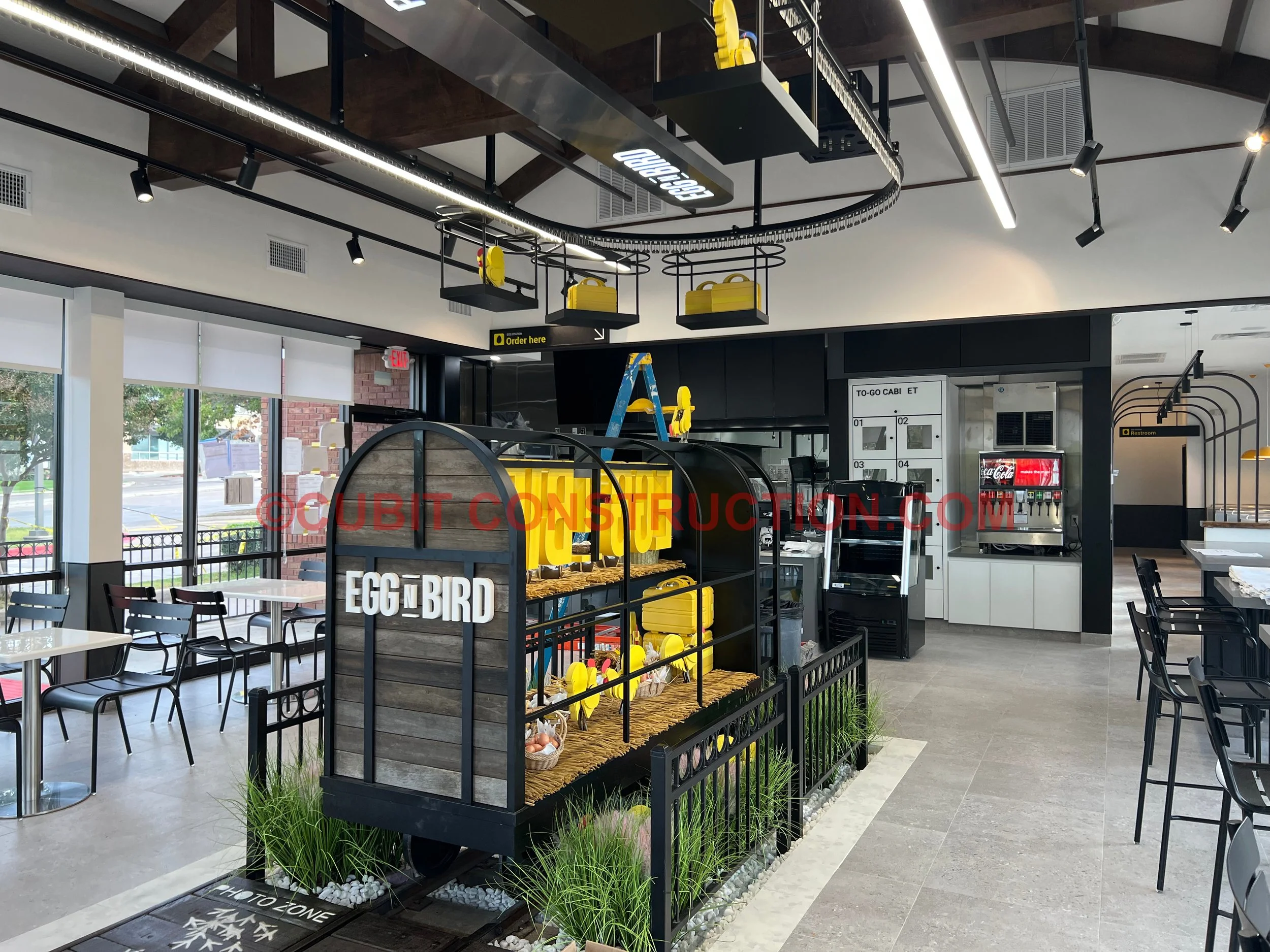 Interior of a modern fast-food restaurant with a black and yellow themed display titled "Egg & Bird" featuring a small train with yellow eggs and chicks, seating area with tables and chairs, vending machines, and self-serve coffee station.