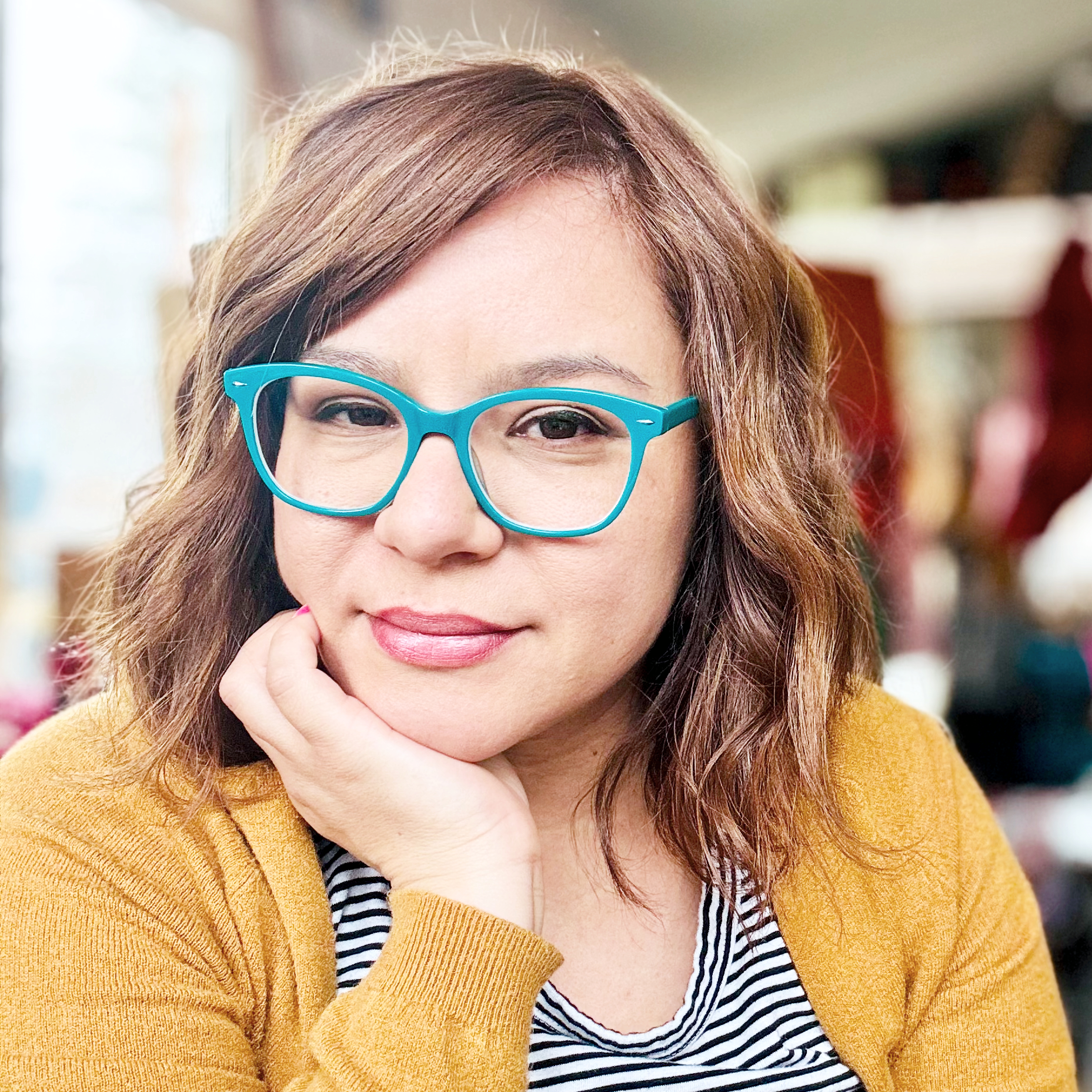 A woman with brown hair, wearing blue glasses, a yellow sweater, and a black and white striped shirt, smiling and resting her chin on her hand.